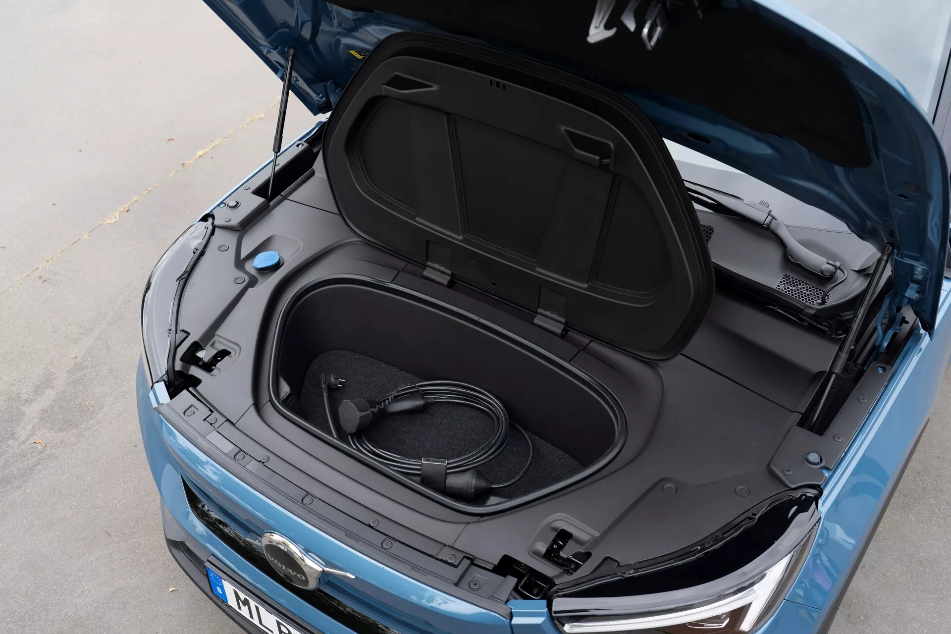 Närbild, vinklad ovanifrån av Volvo C40 2021 (1:a generationen) med frunken öppen, avslöjar tomt förvaringsutrymme och en upprullad laddningskabel, vilket indikerar att det är ett elfordon. Metallisk blå färg, svart frunk-lock inredning med informationsdekaler, delvis synlig tillverkarens logotyp, eleganta strålkastare och synlig främre grill, vilket tyder på ett EV med modern LED-teknik och strömlinjeformad estetik.