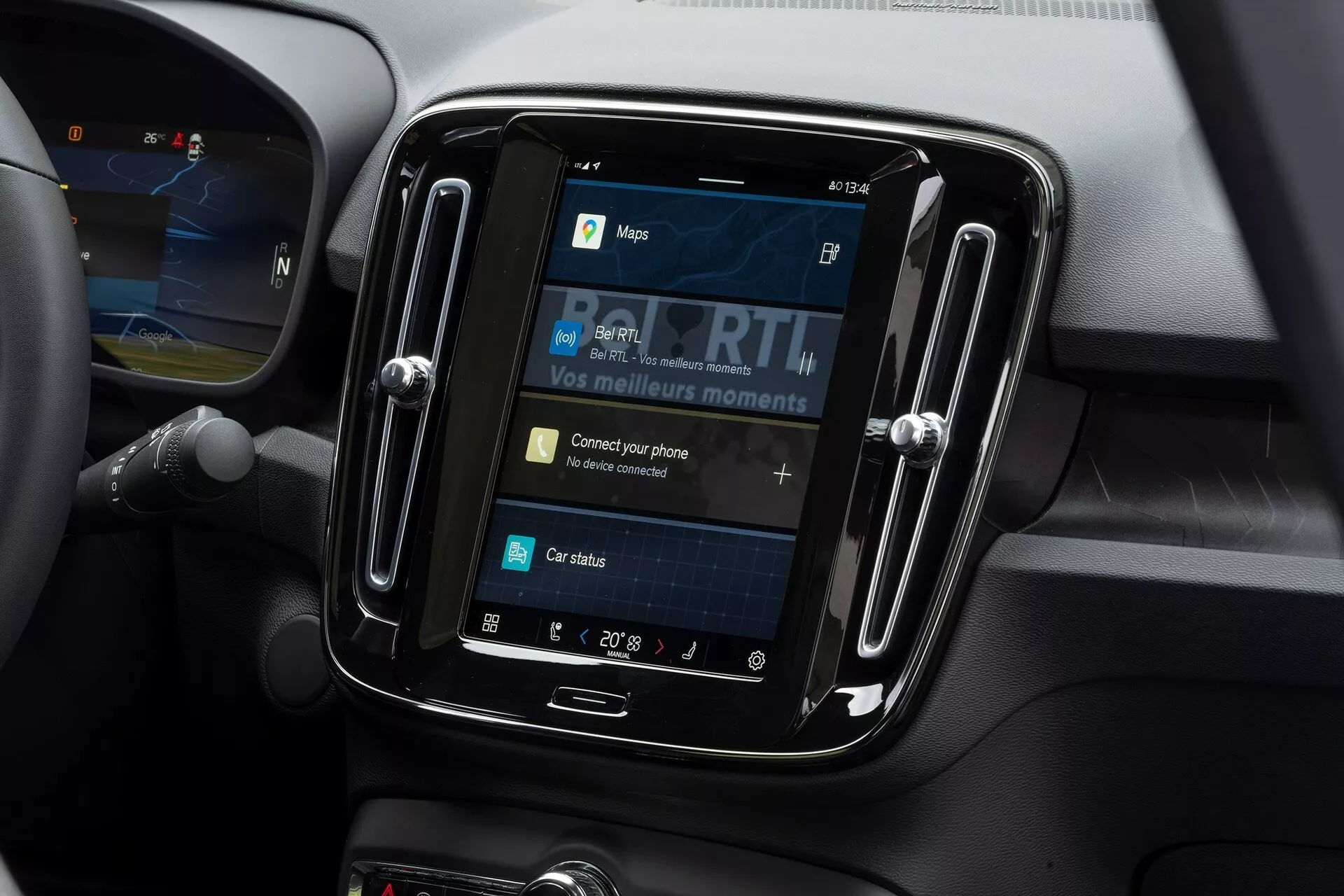 Närbild av infotainmentsystemet inuti en Volvo C40 2021. Bilden, tagen från en lätt nedåtvinklad vinkel, fokuserar på pekskärmen inramad i en glansig, svart kant. På skärmen visas alternativ som kartor, media, telefonanslutning och bilstatus, med 'Anslut din telefon' som ett interaktivt element. Runt infotainmentsystemet finns mjuka material och konturlinjer som antyder ventiler, vilket tyder på en modern bilinteriör. Temperaturinställningen visar 20° Celsius och en del av ratten är synlig till vänster.