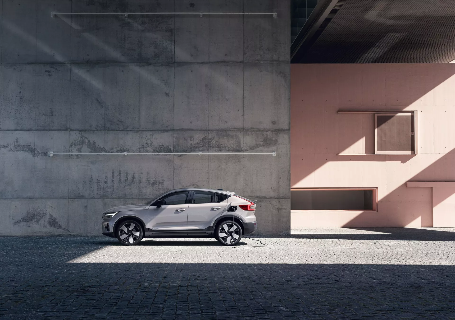 Sidovyprofil av en elegant Volvo C40 SUV från 2021 (1:a generationen) mot en urban bakgrund med betong och persikofärgade väggar. Bilen har en reflekterande vit lackfinish, stiliga lättmetallfälgar, tonade rutor, silvertakräcken och integrerade blinkers i backspeglarna. Fotot framhäver bilens designlinjer och kurvor under naturligt dagsljus, vilket ger en känsla av lyx och sofistikation.