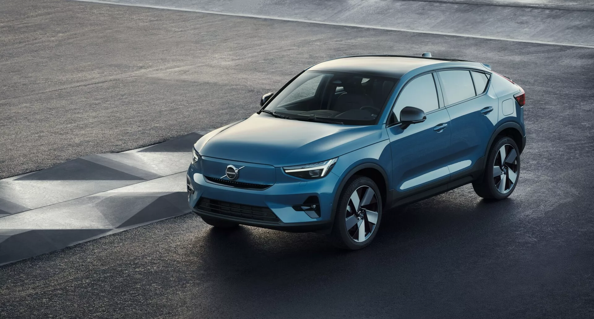 Högvinklat perspektiv av en modern SUV Volvo C40 2021 (1:a generationen) parkerad på en asfalterad yta, visande en trekvartsfönster framifrån. Den eleganta främre delen, troligen karaktäristisk för elektriska fordon med minimala luftintag, förenar sig med skarpa LED-strålkastare, en slät sluttande motorhuv och konturerad sidoprofil med stora lättmetallfälgar. Sidobackspeglar med integrerade blinkers, rikligt med fönsteryta med möjlig privacy tint och en försiktigt sluttande taklinje med takrails för nyttofunktioner är märkbara inslag. Den klara vyen fångar fordonets lyxiga design, avancerade aerodynamik och moderna teknologiska element.