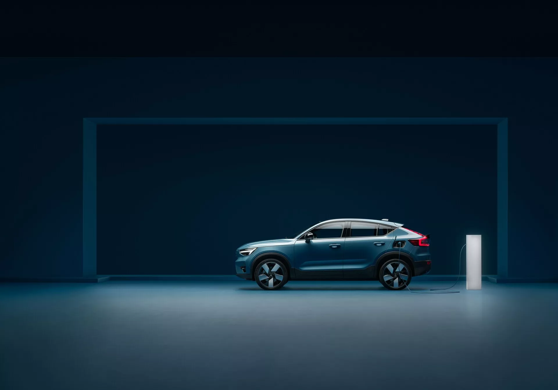 Sidovy av en modern Volvo C40 2021 (första generationen) i en studiouppsättning med reflekterande golv, som visar en elegant design, djupblå metallfärg, sluttande taklinje, stora multifärgade lättmetallfälgar, konturerade linjer, robust konstruktion, integrerade sidospeglar med blinkers, LED-baklyktor och en elektrisk laddningsport.