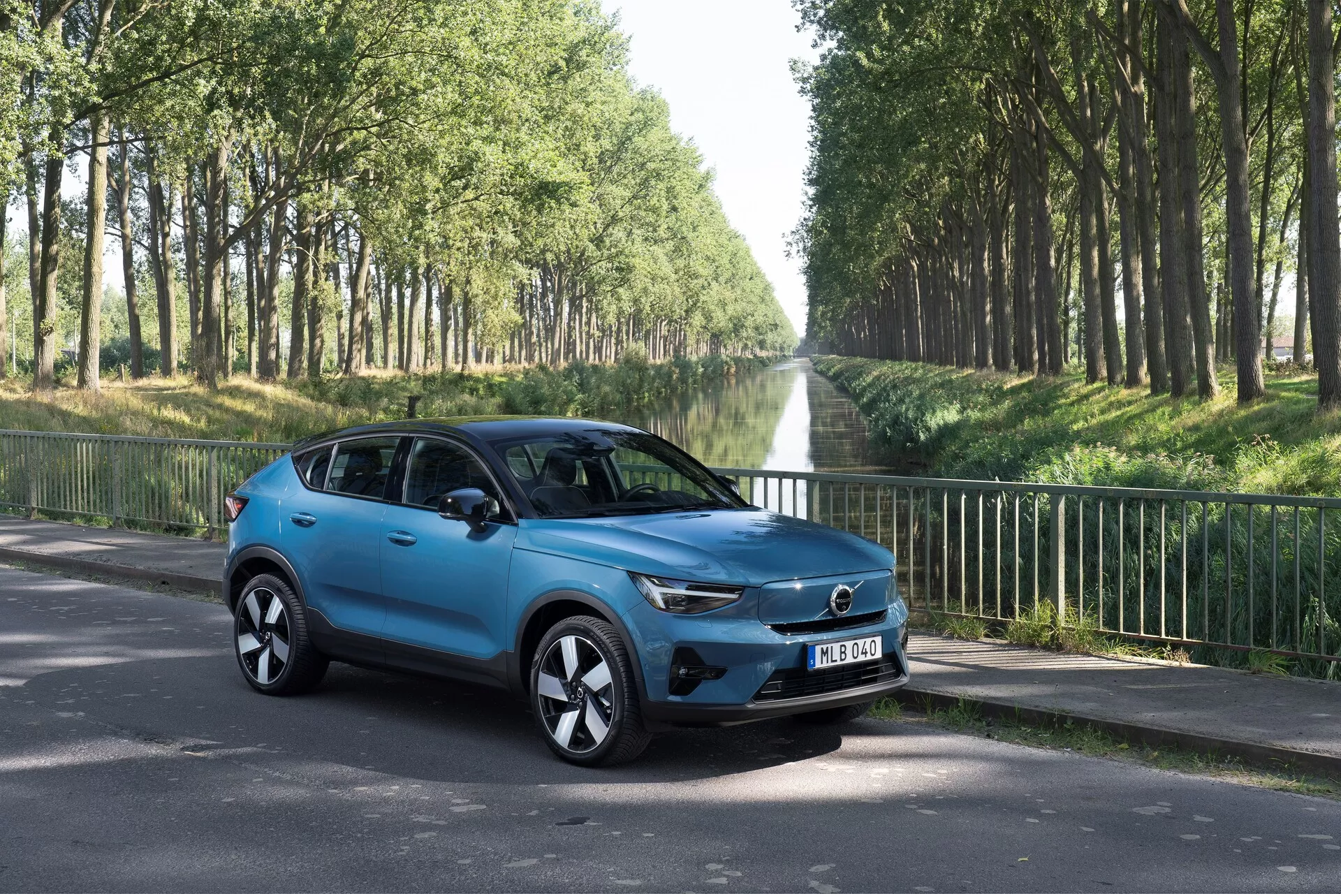 Volvo C40 2021 (Första Generationen) kompakt SUV i trekvarts vy framifrån vänster på en asfaltsväg. Visar moderna designelement som den framträdande grillen med centrerad badge, eleganta LED-strålkastare, stiliga aluminiumfälgar, sidobackspeglar med integrerade blinkers, aerodynamiskt formad kaross med en ljusblå lackfinish, tonade rutor och fyra dörrar. Miljö utomhus med träd längs vägen, fokuserad på estetiska och teknologiska funktioner.