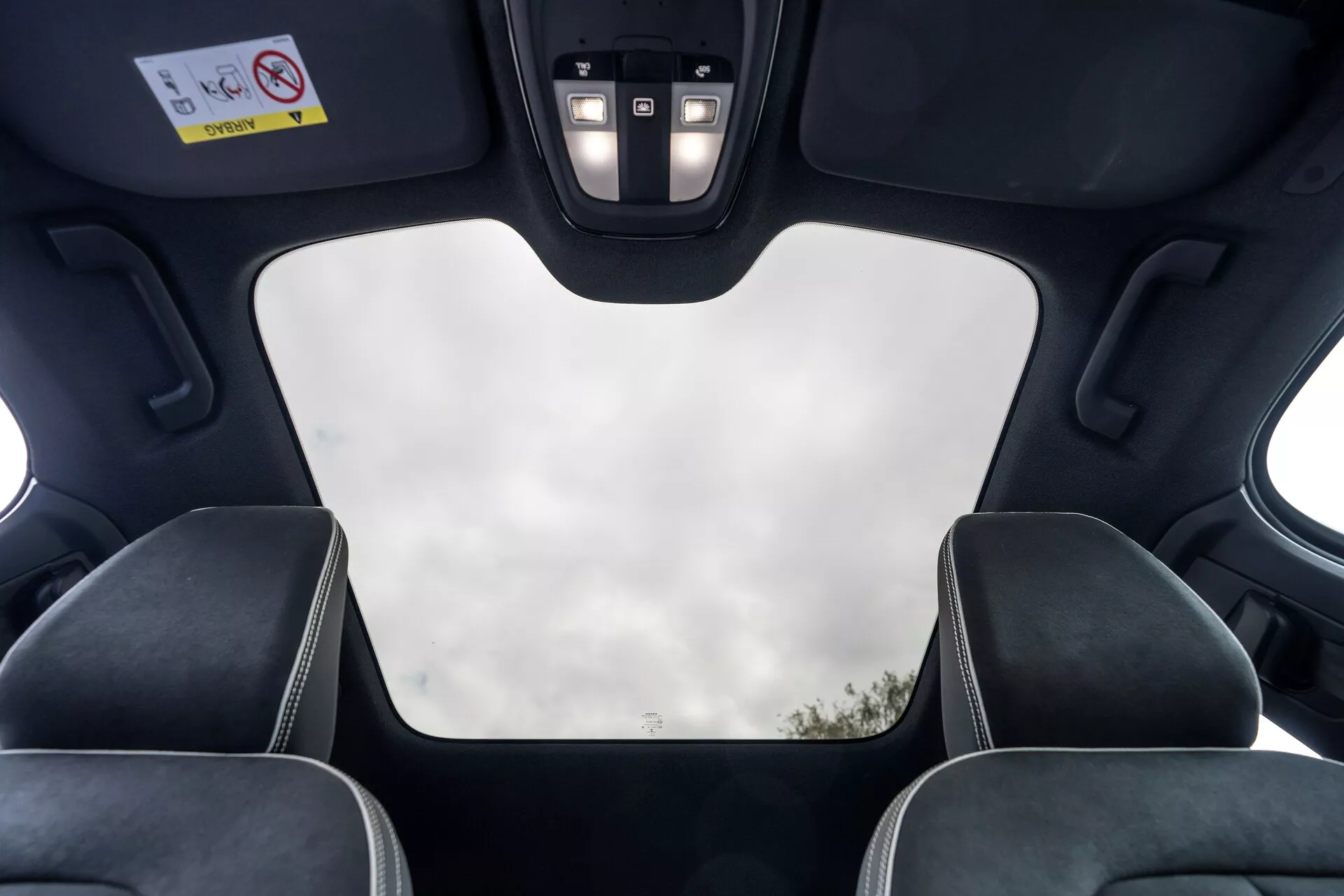 Interiörvy av 2021 Volvo C40 (1:a generationen) från baksätespassagerarens perspektiv, som visar takfunktioner som en stängd panoramatak med klar sikt mot himlen, svart innertak, takmekanismens utskärningar, LED-belysning och kontrollpanel med knappar för olika funktioner. Bilden framhäver också toppdelarna av framsätenas nackstöd med kontrasterande sömmar, vilket betonar fordonets moderna lyxdesign och högkvalitativa funktioner.