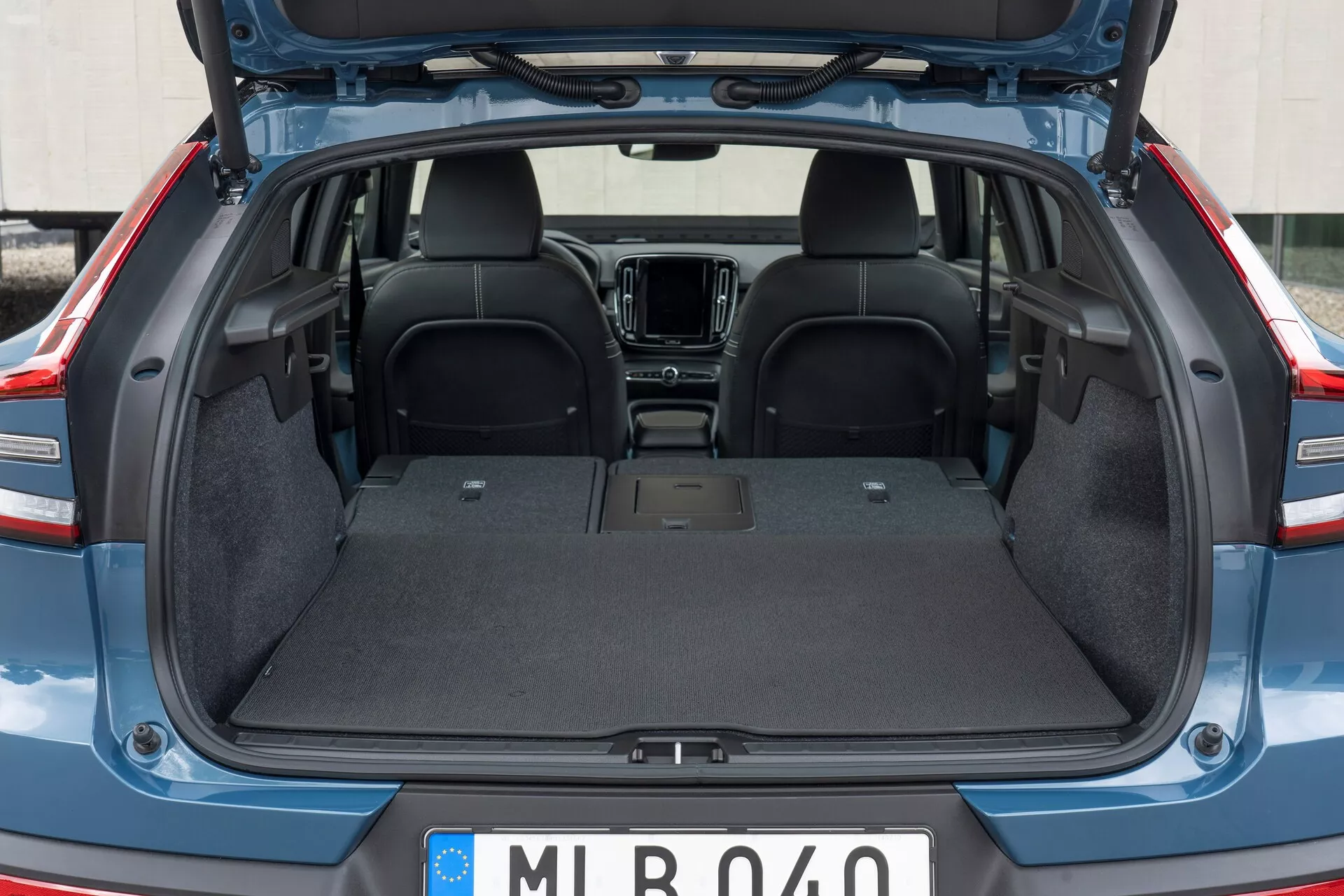 Bakre interiör av Volvo C40 2021 (1:a generationen) med öppen bagagelucka, visar det tomma lastutrymmet med slitstark mattfoder, platt yta och inga synliga intrång. Övre delen av de bakre sätena i mörkt tyg är synlig, med en delvis vy av framsidans interiöra element som mittkonsolen och infotainmentsystemet. Bagageluckan är upphöjd och avslöjar plast-/kompositkanter.