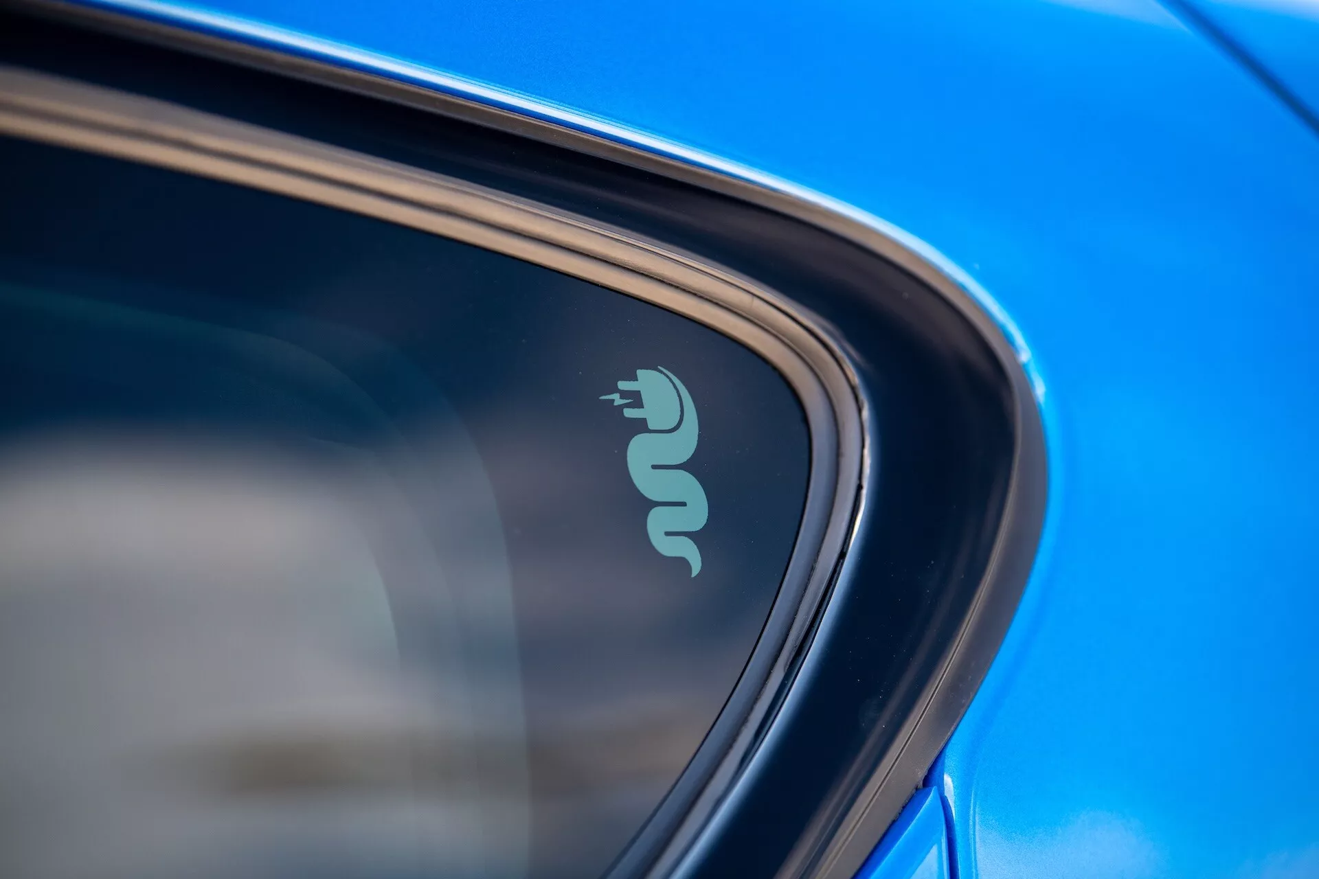 Detaljerad närbild av Alfa Romeo Tonale 2022 som visar teknikmärke som indikerar elektrisk eller hybrid kapacitet nära bilens fönster, framhävd av reflekterande blå färg och slank design i naturligt dagsljus
