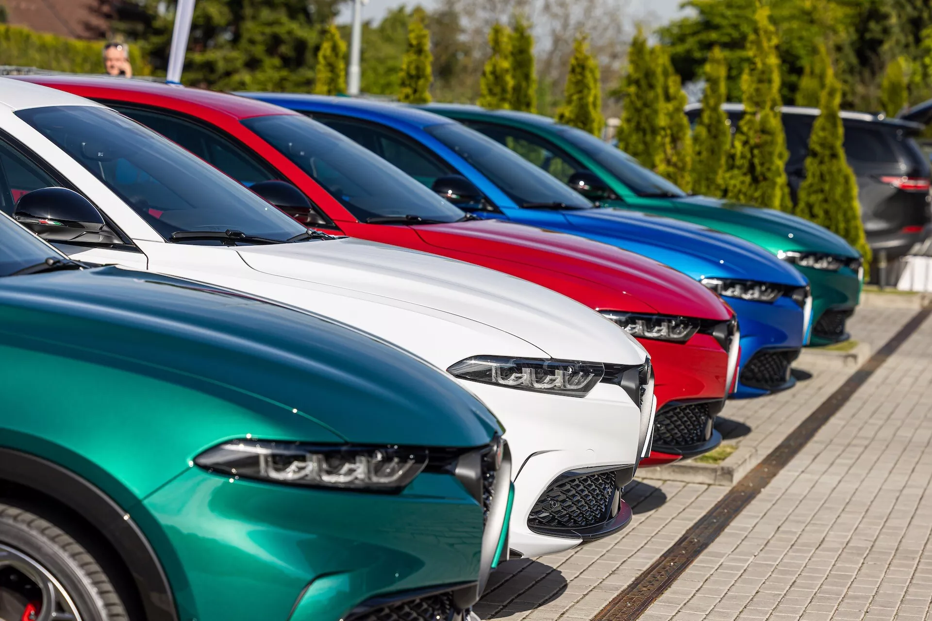 Rad av Alfa Romeo Tonale 2022-modeller parkerade diagonalt som visar fronten från ett tre kvarters perspektiv med fokus på LED-strålkastare, varierande grilldesigns, glansiga färgfinishar och legeringshjul under starkt solljus med bakgrund av grönska