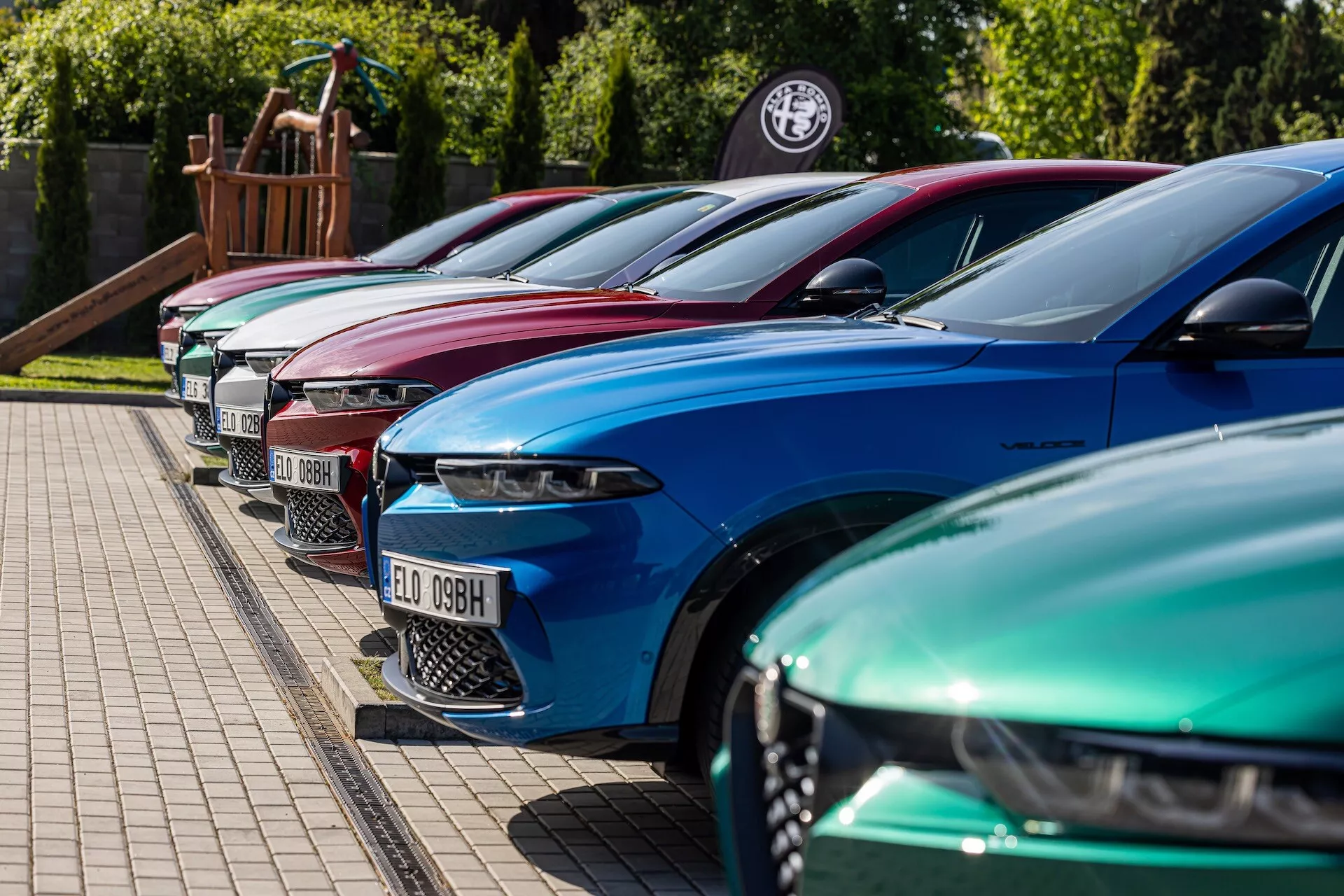 2022 års Alfa-Romeo Tonale-uppställning i livliga färger som visar lyxig design med avancerade LED-strålkastare, parkerade i diagonalt formation med fokus på främre vänster sida från en låg till medium höjdvinkel på ett asfalterat område intill en gräsbrädd, möjligen hos en återförsäljare eller vid ett bilentusiast-evenemang