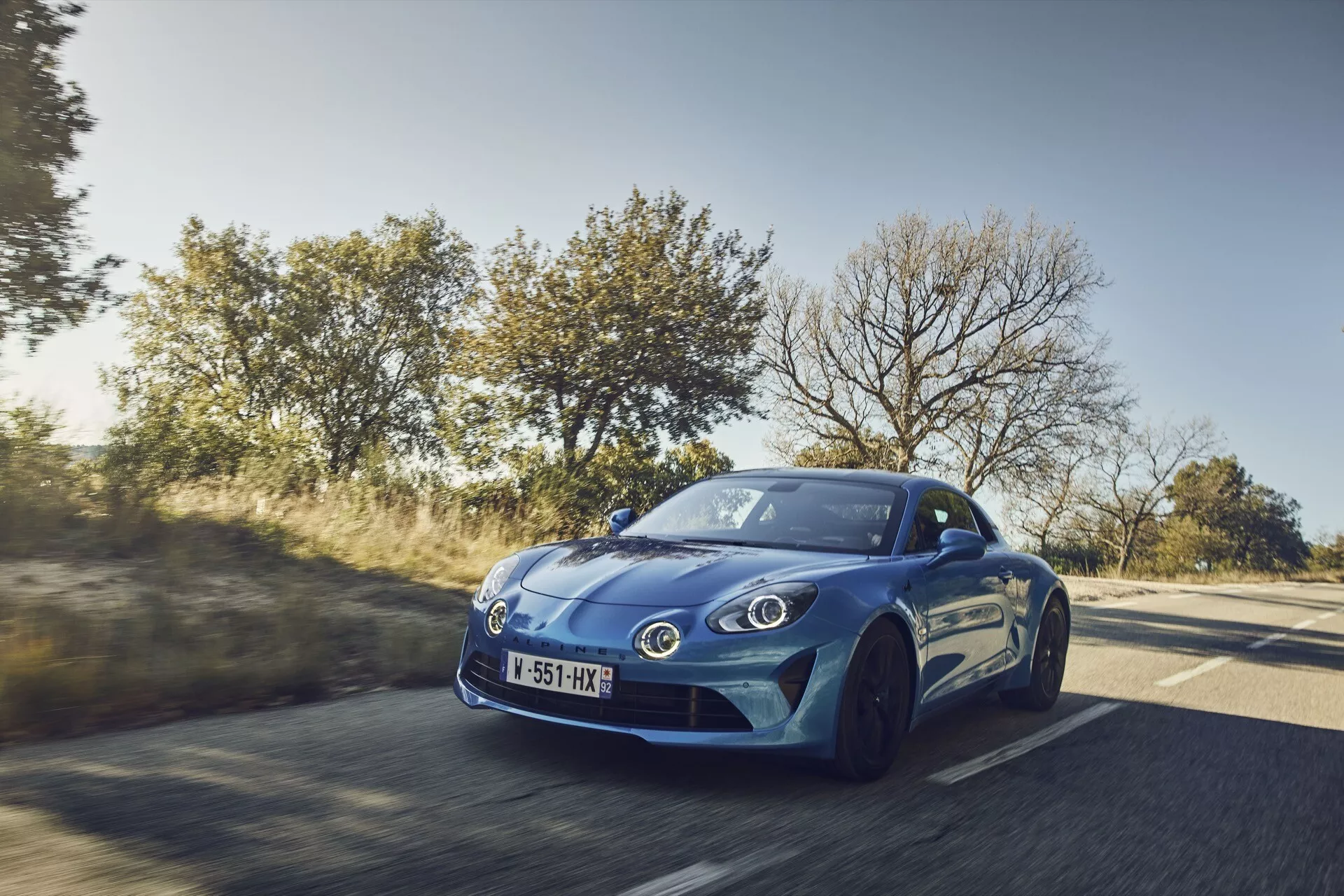 Dynamisk trekvarts frontvy av en blå 2017 Alpine A110 sportscoupé i rörelse på en landsväg, som visar upp aerodynamisk design, moderna strålkastare och sportig hjuluppsättning