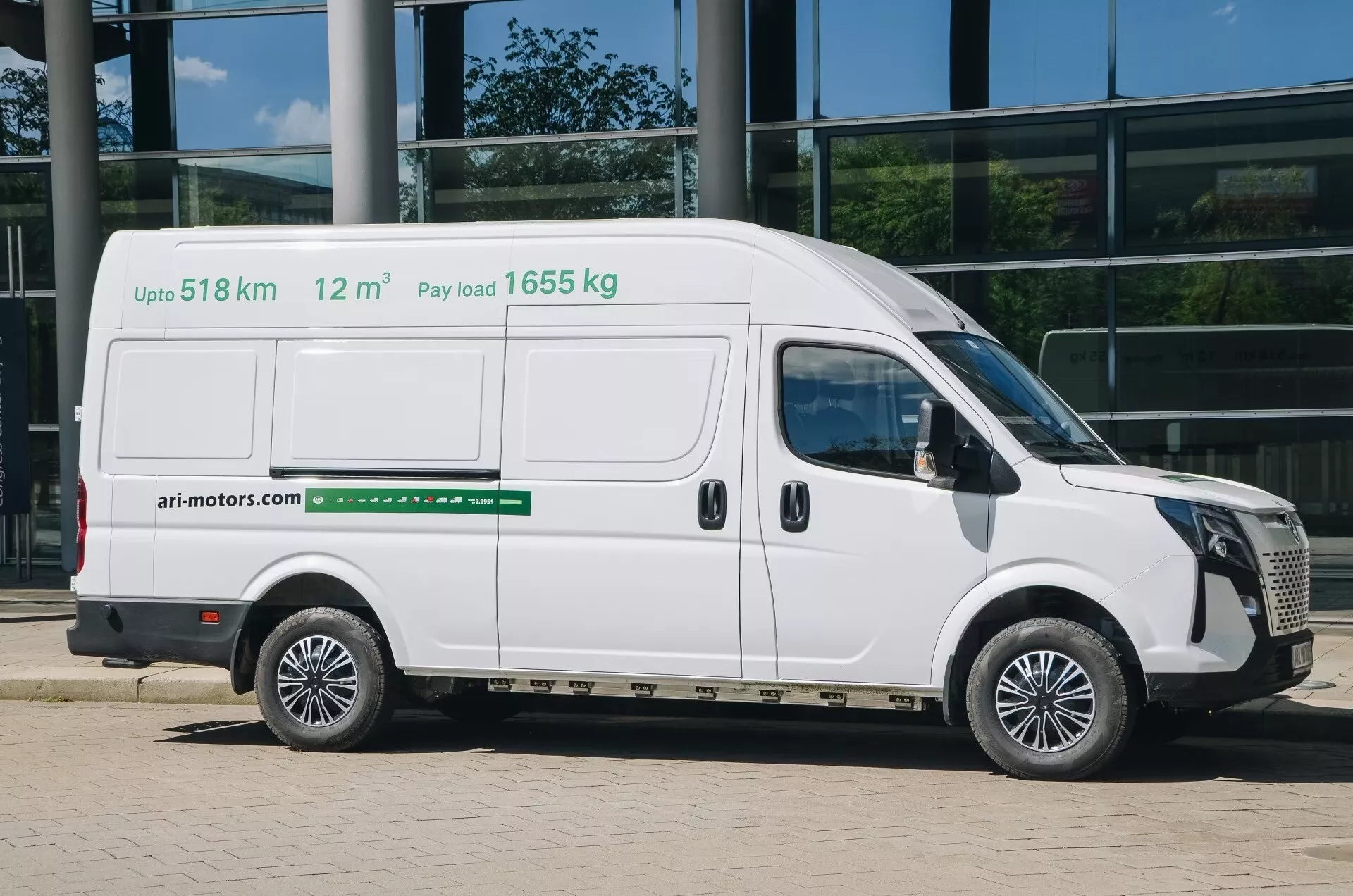 En vit skåpbil för kommersiellt bruk, ari-motors 1710 2024 (1:a generationen), ses från sidan i en detaljerad, professionell bild. Bilden fångar hela passagerarsidan och visar en skjutdörr och två fönster på den övre halvan av fordonet. Text på sidan av skåpbilen anger nyckelfunktioner: 'Upp till 518 km' räckvidd, 'Lastkapacitet 1655 kg' och '12 m³' lastvolym. Detta kan tyda på att skåpbilen är elektrisk eller använder alternativt bränsle. Med en glasväggsbyggnad som bakgrund är fotot taget i dagsljus med skuggor till höger, och framhäver skåpbilens moderna design och kommersiella användning.