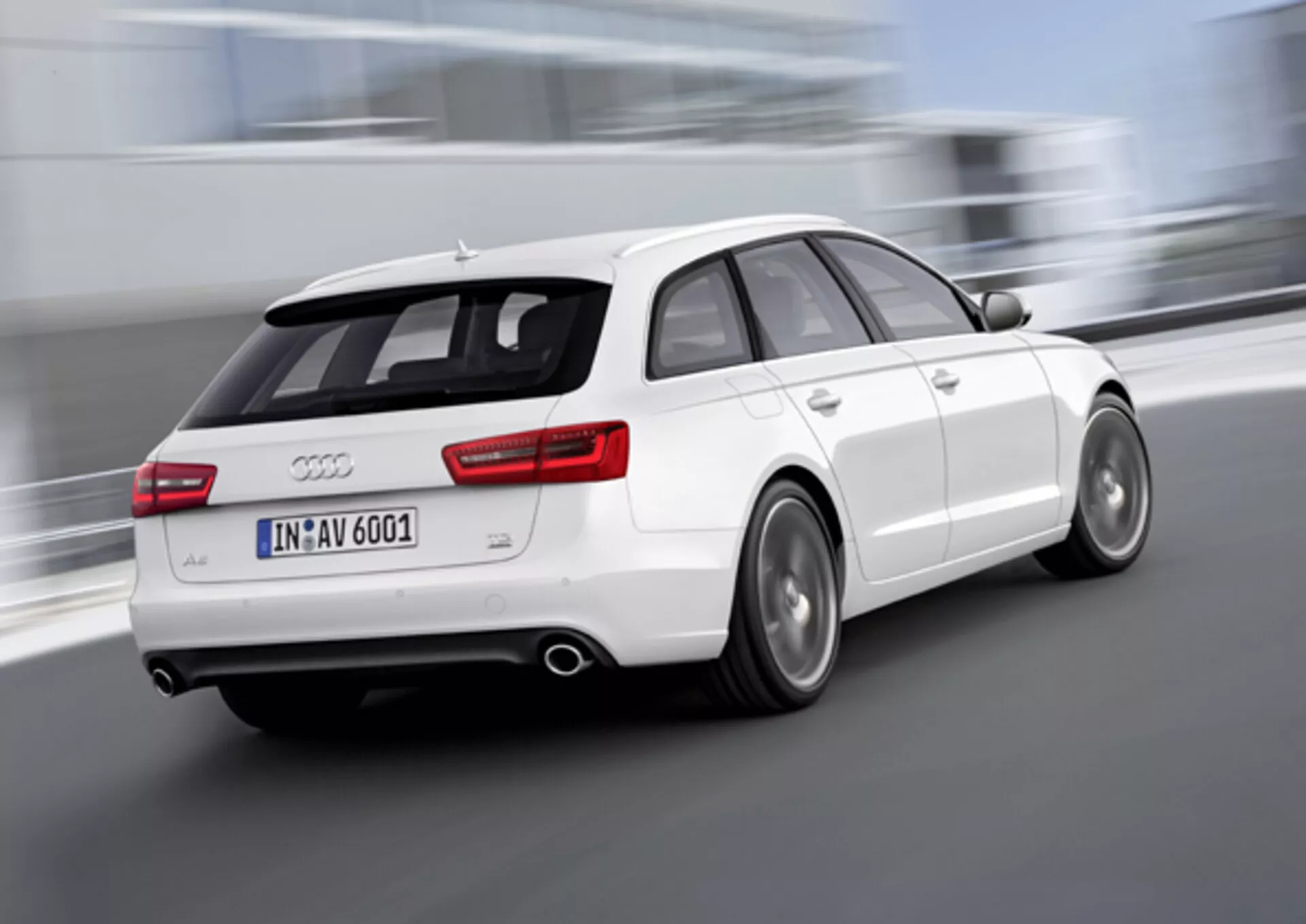 Dynamisk trekvarts bakifrån-vy av en vit Audi A6 Avant från 2011 i rörelse, med suddiga hjul som indikerar hastighet, dubbla avgassystem, legeringshjul med synliga skivbromsar, och en takmonterad spoiler för förbättrad aerodynamik, tagen utomhus.