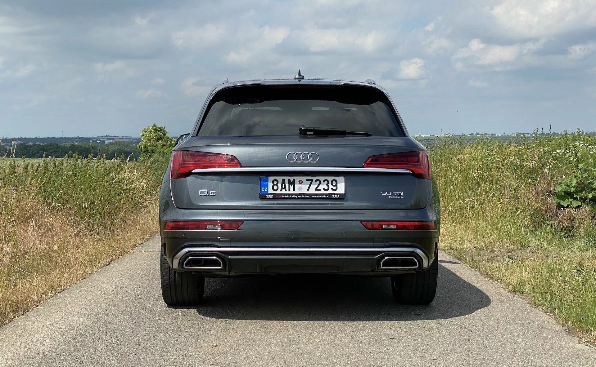 Bakvy från en modern Audi Q5 2020 Sportback SUV som framhäver symmetrin i dess design med distinkta LED-baklyktor, en bakre stötfångare med metalliska accenter och integrerade dubbla avgasrör samt en kromlist ovanför registreringsskylten. Bilen står på en smal trottoar i en landsbygdsmiljö med fält och molnig himmel, som visar dess lyxiga och sportiga egenskaper inklusive parkeringssensorer och en takspoiler.