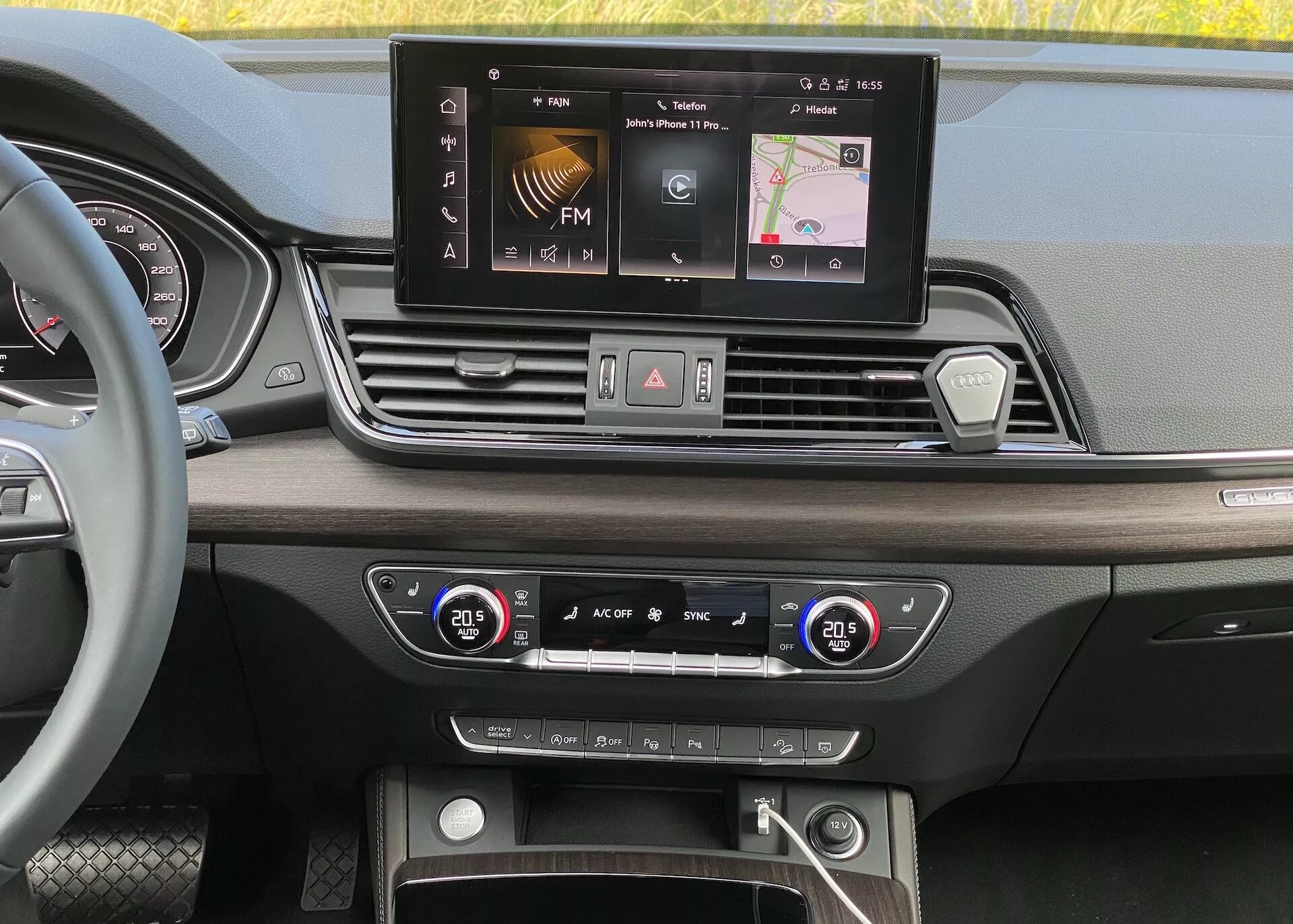 Interiörvy av instrumentpanelen på Audi Q5 2020 Sportback från ögonhöjdsperspektiv. Bilden fokuserar på bredbilds-infotainmentskärmen med alternativ som FM-radio, navigation och fordonsinställningar. Nedanför skärmen finns fyra symmetriskt placerade luftventiler samt en central konfiguration av knappar och vred för varningsblinkers, avfrostning och snabbåtkomstinställningar. Systemet för tvåzons-klimatkontroll med digitala temperaturvisningar är inställt på 20,5 grader Celsius för både förare och passagerare. Den nedre delen visar ett förvaringsutrymme med möjliga USB-portar, en hjälpingång och en start/stopp-knapp för nyckelfri startteknik. De lyxiga materialen och designen inkluderar träimitationer och polerade metallaccenter.