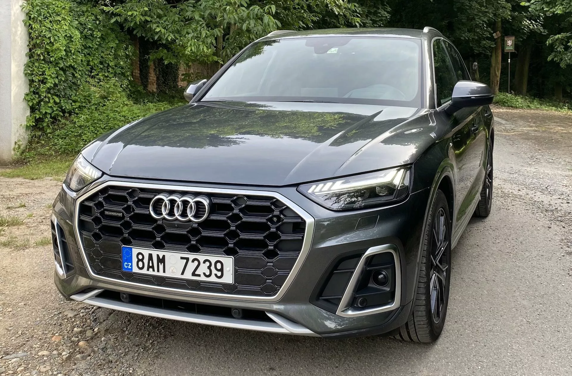 Trekvarts framsida av en Audi Q5 Sportback 2020 som visar vänster främre sida med den karakteristiska hexagonala bikakegrillen, Audi-logotypen, eleganta vinklade LED-strålkastare och stora luftintag på den nedre stötfångaren. Bilen är parkerad på en väg omgiven av träd som reflekterar Blixtfärgen. Den skulpterade motorhuven med aerodynamiska linjer mot emblemet och den främre flerspakade lättmetallfälgen betonar dess sportiga, moderna design.