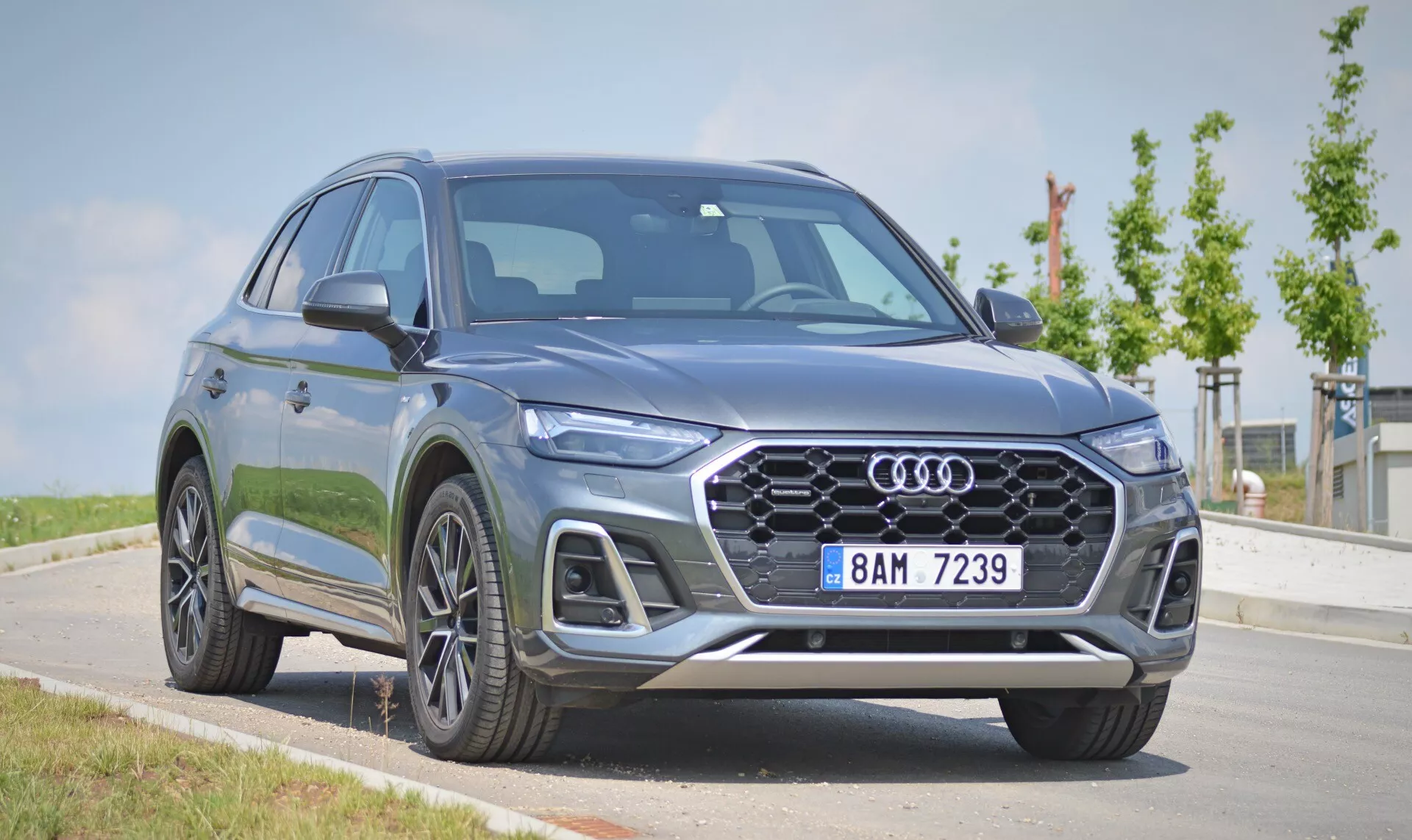 Bilden visar en Audi Q5 Sportback 2020, en modern lyxcrossover-SUV. Fångad från en låg främre trekvartsvinkel, framhäver den den framträdande hexagonala frontgrillen, LED-strålkastare med distinkt dagljussignatur, legeringshjul och skulpterade karosslinjer. Fordonet är parkerat på en asfaltyta med klar himmel och grönska i bakgrunden, vilket ger en detaljerad vy av dess metalliska grå eller blå finish och premiumdesignfunktioner.