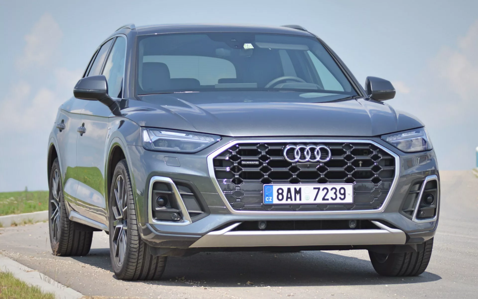 Modern Audi Q5 2020 (Q5 Sportback) SUV fotograferad från en låg trekvartsvinkel framifrån, med fokus på fronten, hexagonal grill med fyraringsemblem, eleganta LED-strålkastare och en del av sidoprofilen. Den stora, mönstrade grillen, integrerade dimljus, luftintag och silverdekorationer på främre stötfångaren bidrar till både estetisk och funktionell charm. Avancerad belysning och förarassistanssystem antyds av den skarpa strålkastardesignen och grillkonfigurationen. Stora lättmetallfälgar och dynamiska karosslinjer indikerar en premium, robust närvaro. Stillastående på en väg med en klar himmel i bakgrunden, framhäver fordons främre detaljer och aggressiva stilindikationer.