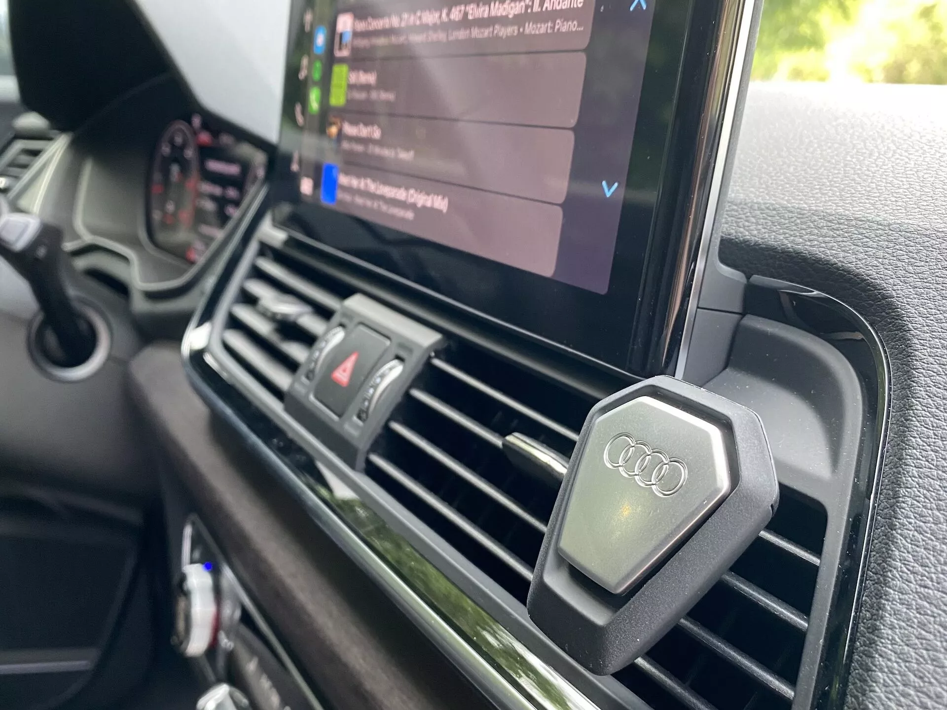 Närbild från förarsidan som visar den centrala instrumentpanelen i en Audi Q5 Sportback 2020. Fokus är på de centrala luftventilerna och infotainmentskärmen som visar en medieuppspelning med en lista över ljudfiler, vilket framhäver fordonets multimediakapacitet. Luftventilerna är rektangulära med en metallfinish och har knoppar med Audi-logotypen. Instrumentpanelen har en texturerad yta av högsta kvalitet i linje med Audis lyxpositionering, och det finns inga synliga manuella luftkonditioneringskontroller, vilket tyder på ett automatiskt klimatsystem.
