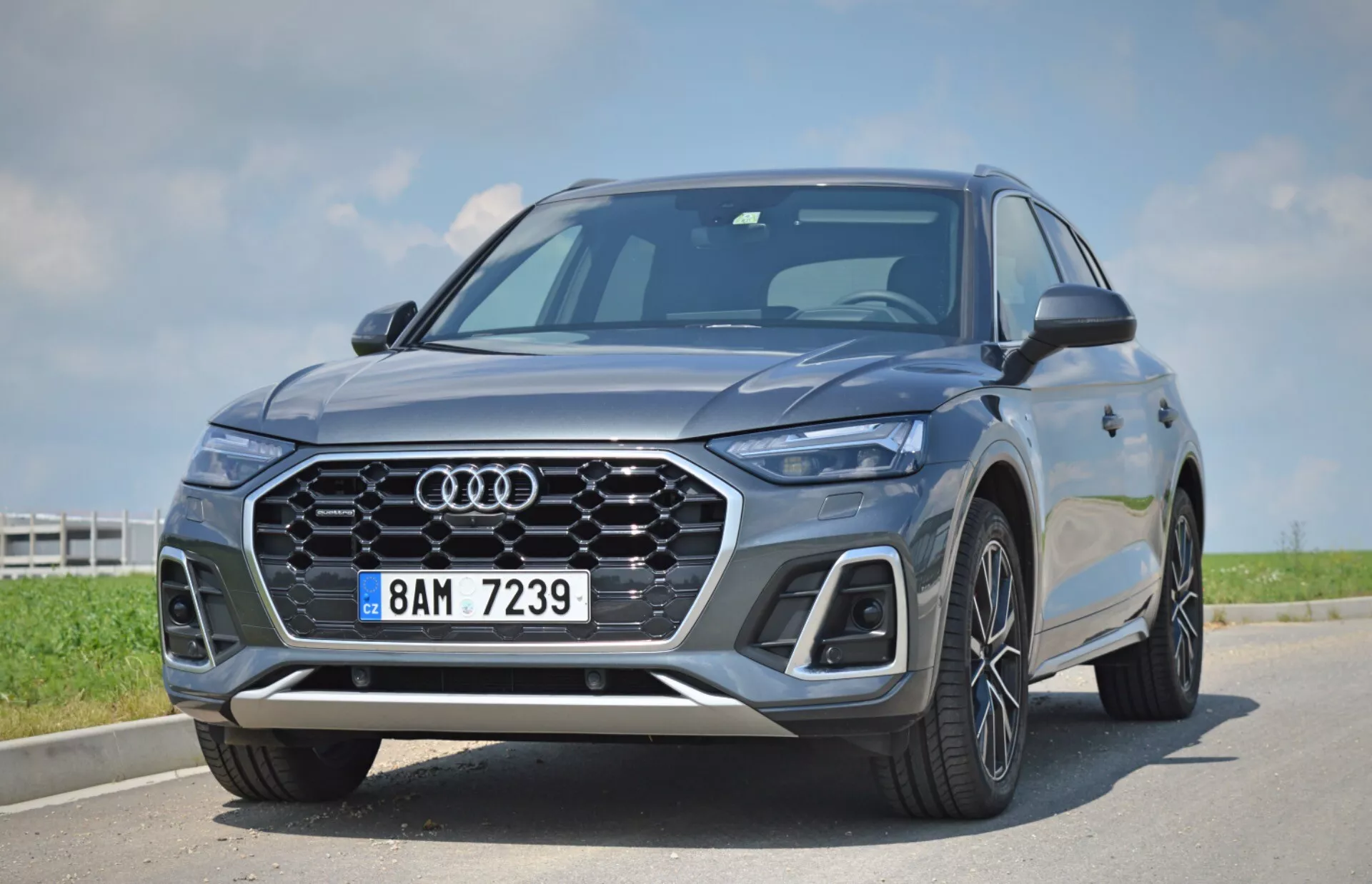 Bilden visar en modern 2020 Audi Q5 Sportback SUV som fångats från en låg och nära vinkel, vilket framhäver fordonets front och grill. Framträdande detaljer inkluderar Audis ikoniska fyraringade emblem, ett distinkt enkelramgsgrill med bikakedesign, eleganta LED-strålkastare med karakteristiska dagsljuslampor, integrerade sensorer i främre stötfångaren som indikerar avancerade förarassistanssystem, stora flerspoks aluminiumhjul, skarpa karosslinjer och aerodynamiska sidospeglar. Fordonet är parkerat under en klar himmel, vilket betonar dess lyxiga och tekniskt avancerade design.