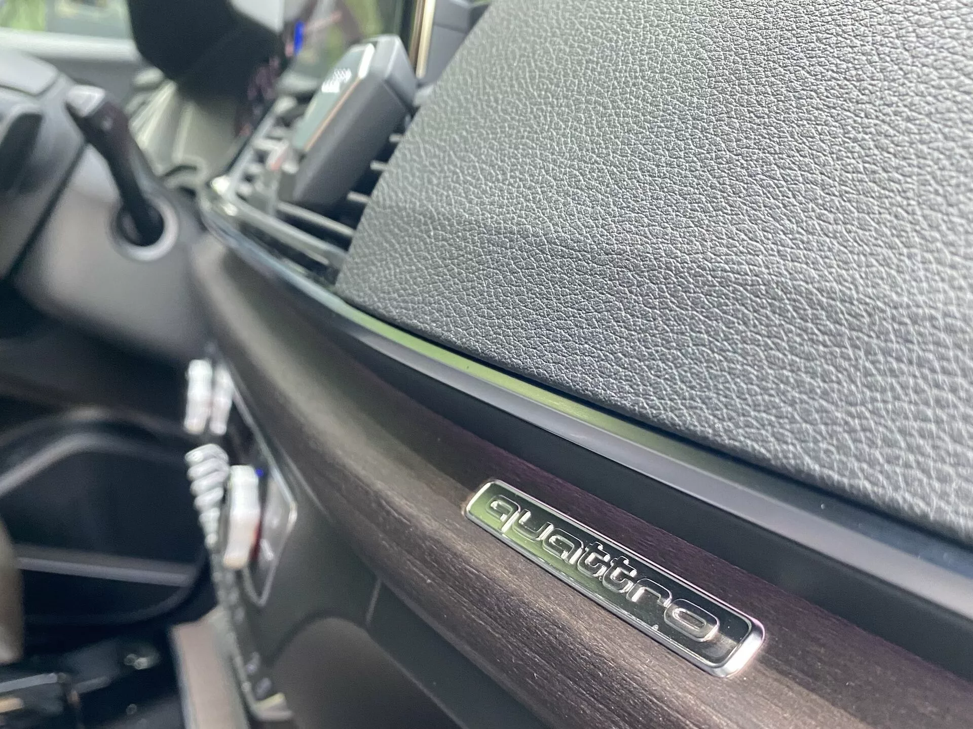 Närbild av interiören i Audi Q5 2020 Sportback, fokuserad på 'QUATTRO'-emblemet i ögonhöjd, vilket framhäver Audis fyrhjulsdriftsystem. Med en glansig finish på emblemet och kontrasterande texturerade material är bilden tagen från en sned vinkel med grunt skärpedjup, vilket visar detaljer av instrumentbrädan och den centrala kontrollpanelen i en suddig bakgrund.