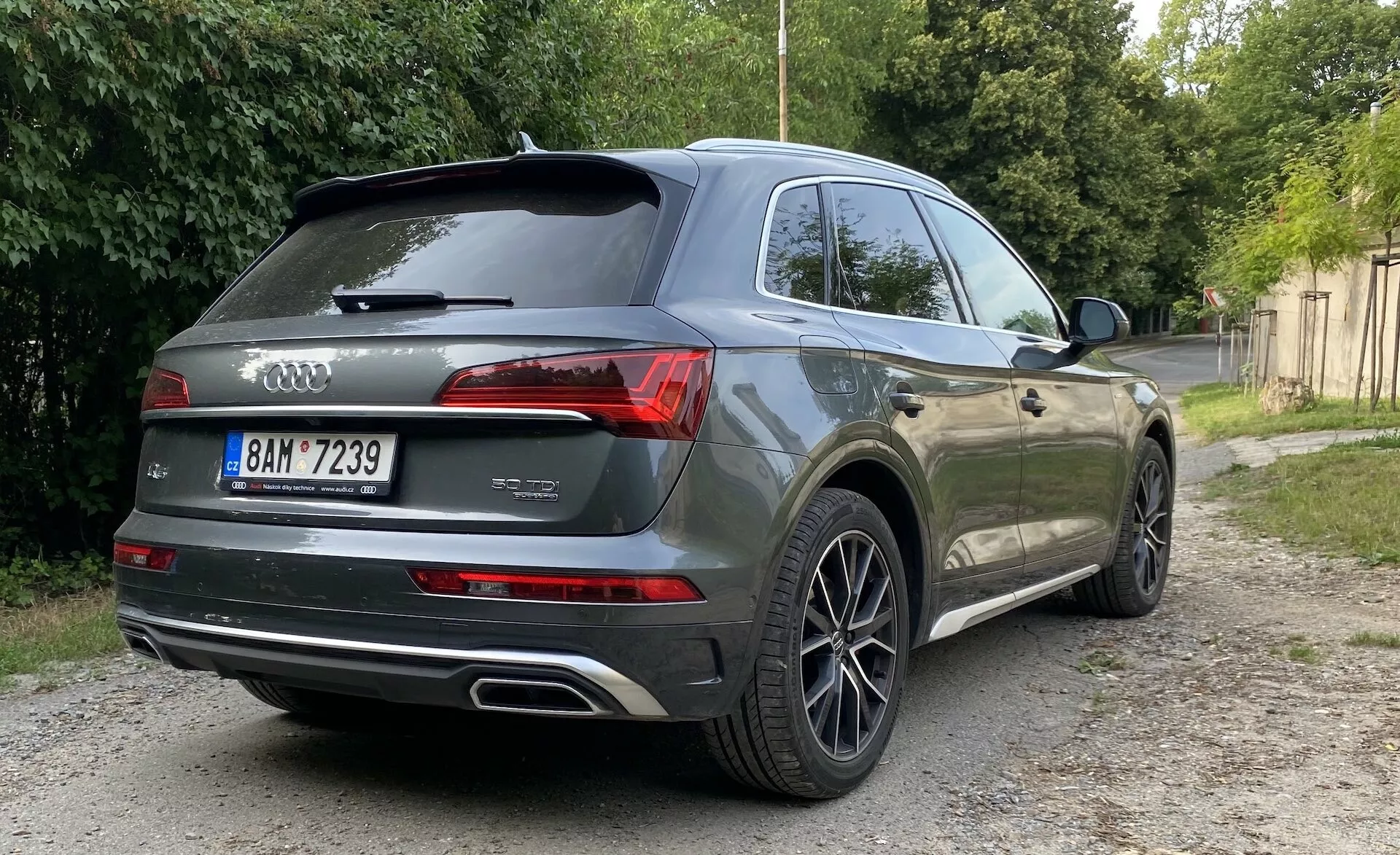 Bakåtvinklad vy av en grå Audi Q5 Sportback 2020 SUV parkerad på en grusbelagd yta bredvid grön vegetation. Bilden fokuserar på bilens bakre och sidovy, visar moderna LED-bakljus, dubbla avgasrör, Audi ringarna-logga, modell 'Q7' och variant '45 TDI' märken, flerspinskade lättmetallfälgar, privata rutor, takspoiler och eventuella parkeringssensorer eller kamera. Naturlig belysning med mjuka skuggor antyder soluppgång eller solnedgång.