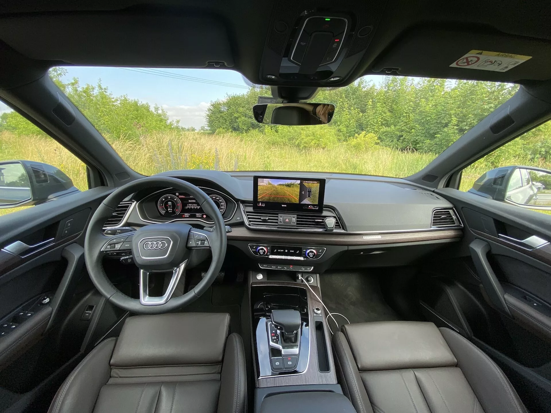 Interiör av 2020 Audi Q5 Sportback fotograferad från förarsätets perspektiv, som visar en lyxig och tekniskt avancerad instrumentpanel. Bilden lyfter fram svarta lädersäten, en mittkonsol med ett läderarmstöd, en automatisk växelspak omgiven av kontrollknappar, en horisontellt orienterad infotainment-pekskärm med GPS-navigering, dubbelzons klimatanläggning, en digital förardisplay bakom en Audi-logotypmärkta ratt med inbyggda kontroller. Designen utmärker sig med minimalism och borstad metallaccent, vilket ger fullständig synlighet genom bilens fönster och stolpe, vilket indikerar att fordonet är parkerat utomhus.