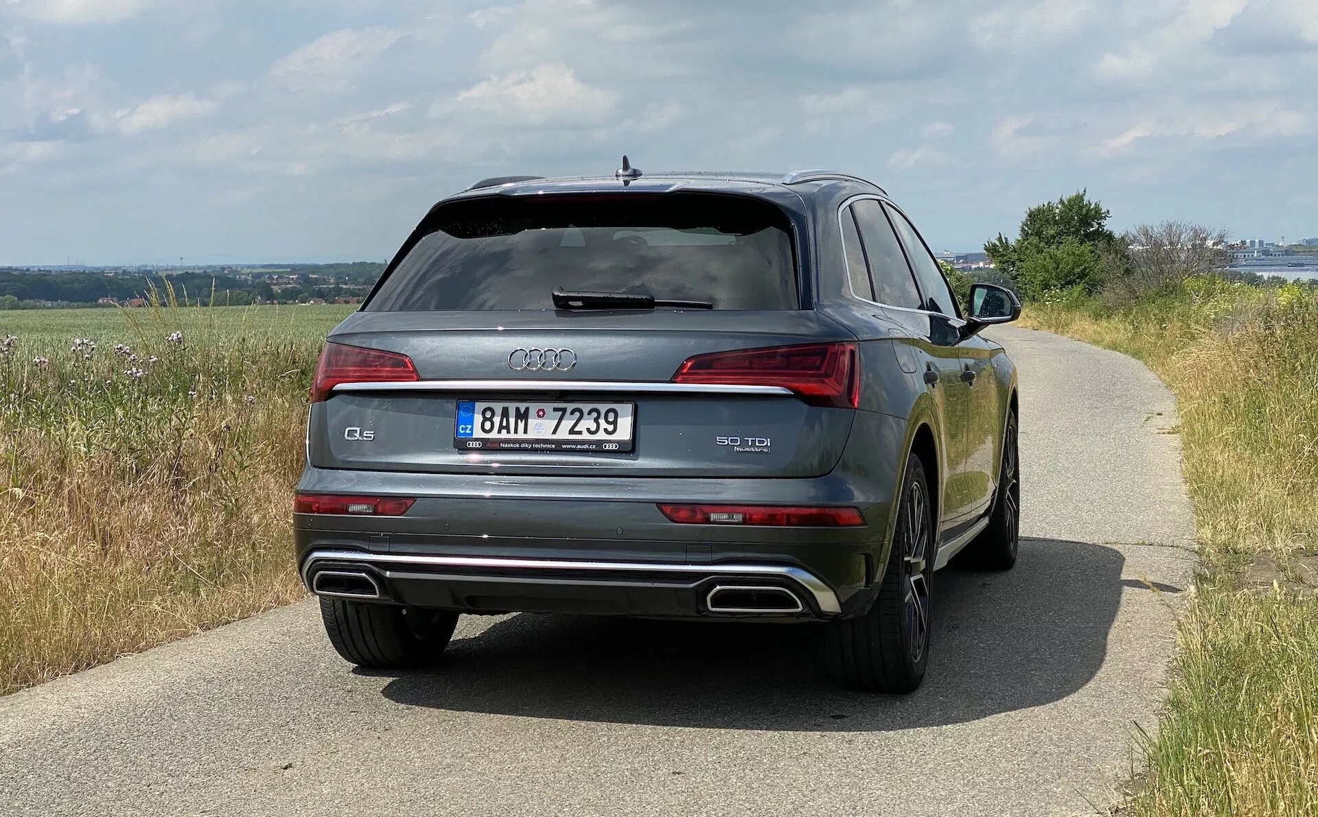 Bakre vy av en 2020 Audi Q5 Sportback som visar bilens bredd inklusive bakljus, bakruta och dubbla avgasrör. Bilen, belägen på en väg i dagsljus som visar en lantlig omgivning, har en blank lack som reflekterar omgivningen. Designdetaljer som Audi-ringar, modellbeteckning och registreringsskylt är tydligt synliga. Tekniska funktioner som LED-ljus, parkeringssensorer och en avfrostad bakruta är också uppenbara.