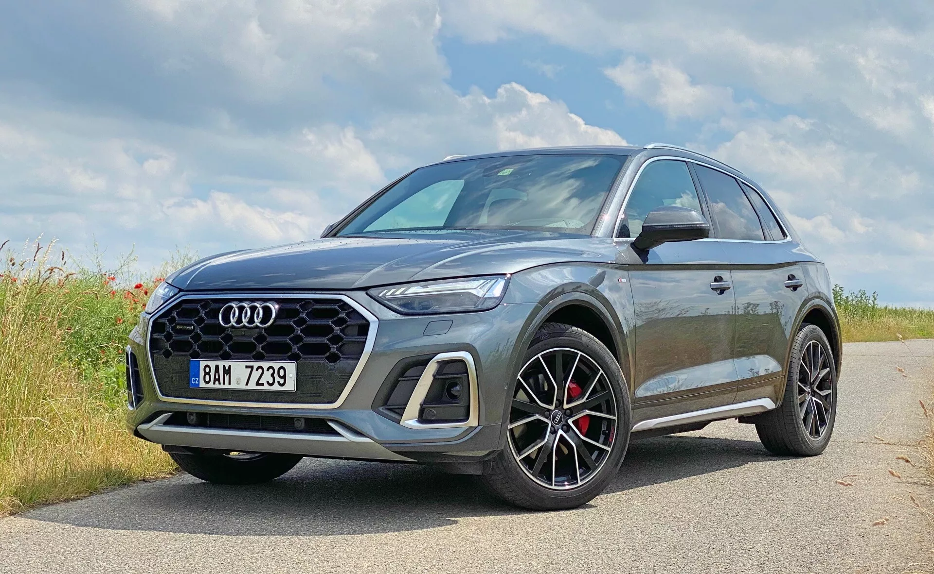 Bild av en modern Audi Q5 Sportback 2020 SUV parkerad på en asfalterad sidoväg i en naturlig miljö, möjligen en lantlig omgivning. Framhävt i förgrunden är Audis fyraringslogo. Denna närbild i trekvartsvy framifrån betonar fordonets vänstra framsida och delvis profil, med fokus på designelement som den hexagonala nätmönstrade frontgrillen, skarpa vinklade LED-strålkastare och stora, mångekrade hjul med svarta detaljer och röda bromsok. Den metalliska grå färgen reflekterar ljuset och förstärker bilens konturer. Den något förhöjda markfrigången hos SUV:en antyder mångsidighet och lätt terrängkapacitet, och sidospeglarna har en kontrasterande färg som antyder en sportig trimnivå. Bilden fångar det lyxiga och sportiga utseendet hos fordonet med eventuella avancerade teknologier, även om de inte är synliga.