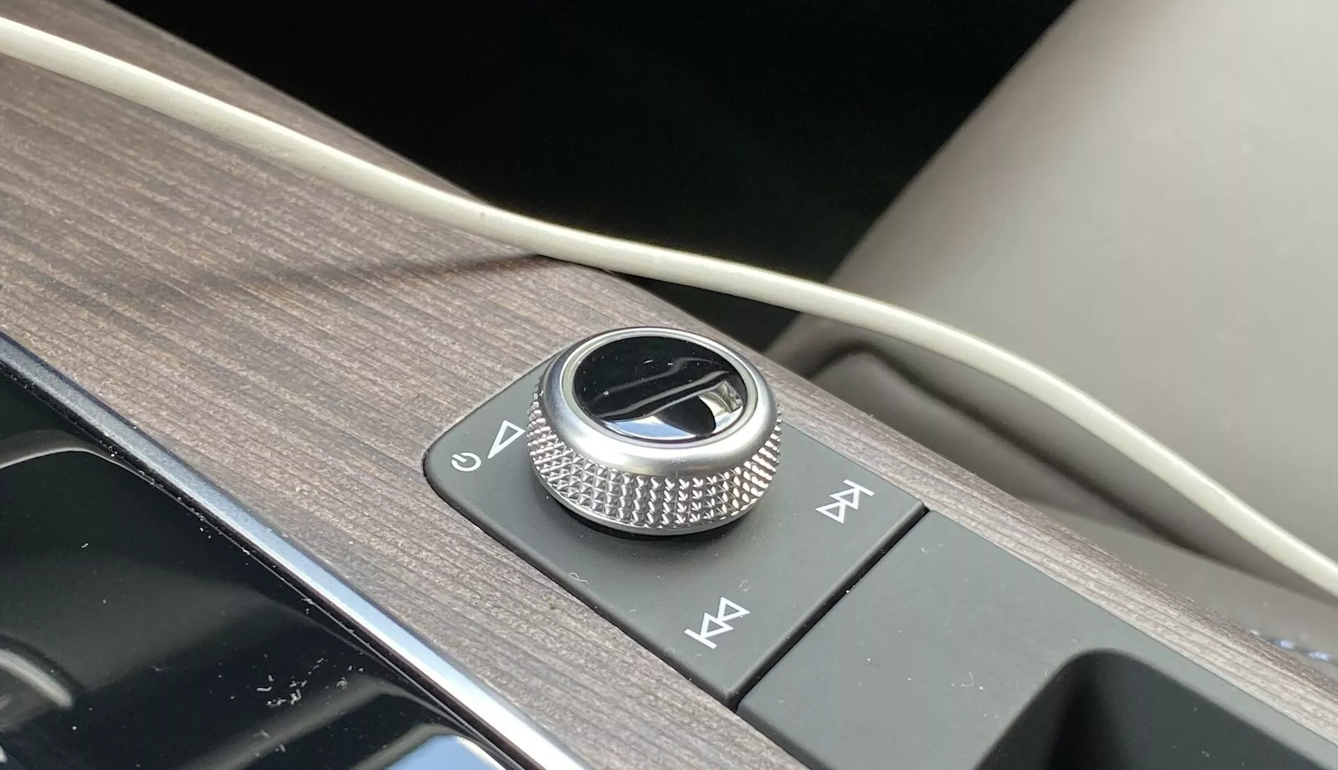 Närbild av mittkonsolen på Audi Q5 2020 Sportback, med en metallisk roterande ratt för infotainmentsystemet omgiven av borstad metall på en glansig svart panel. Bredvid finns två snabbvalsknappar, allt placerat på en yta med trästruktur och en touch av mjukt material som exemplifierar lyxig interiördesign och avancerade infotainmentsystemfunktioner.