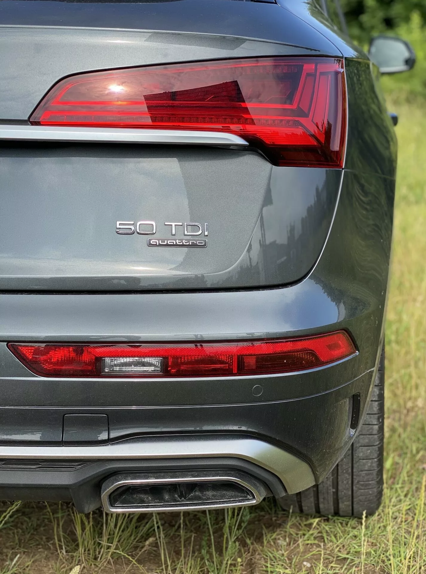 Närbild av Audi Q5 2020 Sportbacks bakre sektion från en bakre trekvartsperspektiv, visar vänster bakljus med LED-element, modellbeteckning '50 TDI' och 'quattro' på bagageluckan, dubbelt avgasrör, metallicgrå lack och en synlig parkeringssensor bak.