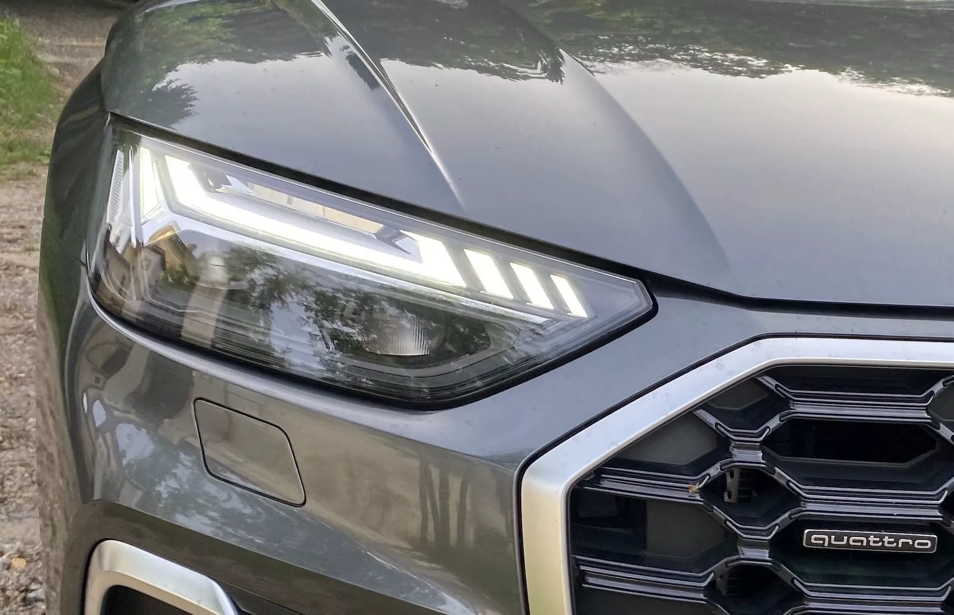 Närbild av vänster framdel på en modern bil som visar en delvis vy inklusive vänster LED-strålkastare, en del av motorhuven och en del av frontgrillen med polygonal design där en 'quattro'-skylt är synlig. Bilden fångar bilens designelement och belysningsteknologi med en metallisk lack, tagen från en nivåperspektiv något uppåtvinklad.