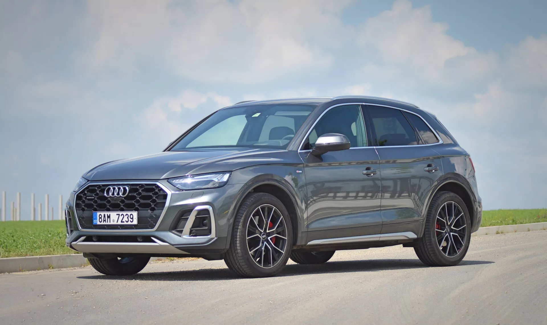 Bild av en modern crossover/SUV, Audi Q5 Sportback 2020 års modell, stationerad på en asfaltyta. Bilen presenteras i en trekvarts framvy som fångar både fram- och sidprofilen. Den framträdande grillen med ett glänsande svart kryssmönster har tillverkarens emblem centralt placerat. De slanka, skarpa LED-strålkastarna och fler-ekrade aluminiumfälgar förbättrar den lyxiga estetik. Den metalliska lacken reflekterar solen, vilket indikerar en premium finish. Sidprofilen visar aerodynamiska karosserilinjer och ytterbackspeglar med integrerade blinkers. Fönstren har en krom trim som kontrasterar med de mörka tonade glasen, vilket kan antyda integritet. Bilen är stationär, placerad något till höger om kamerans centrala axel med skuggor som tyder på att det är antingen morgon eller eftermiddag, under en klar himmel med några få moln i en lantlig eller halv-lantlig miljö.