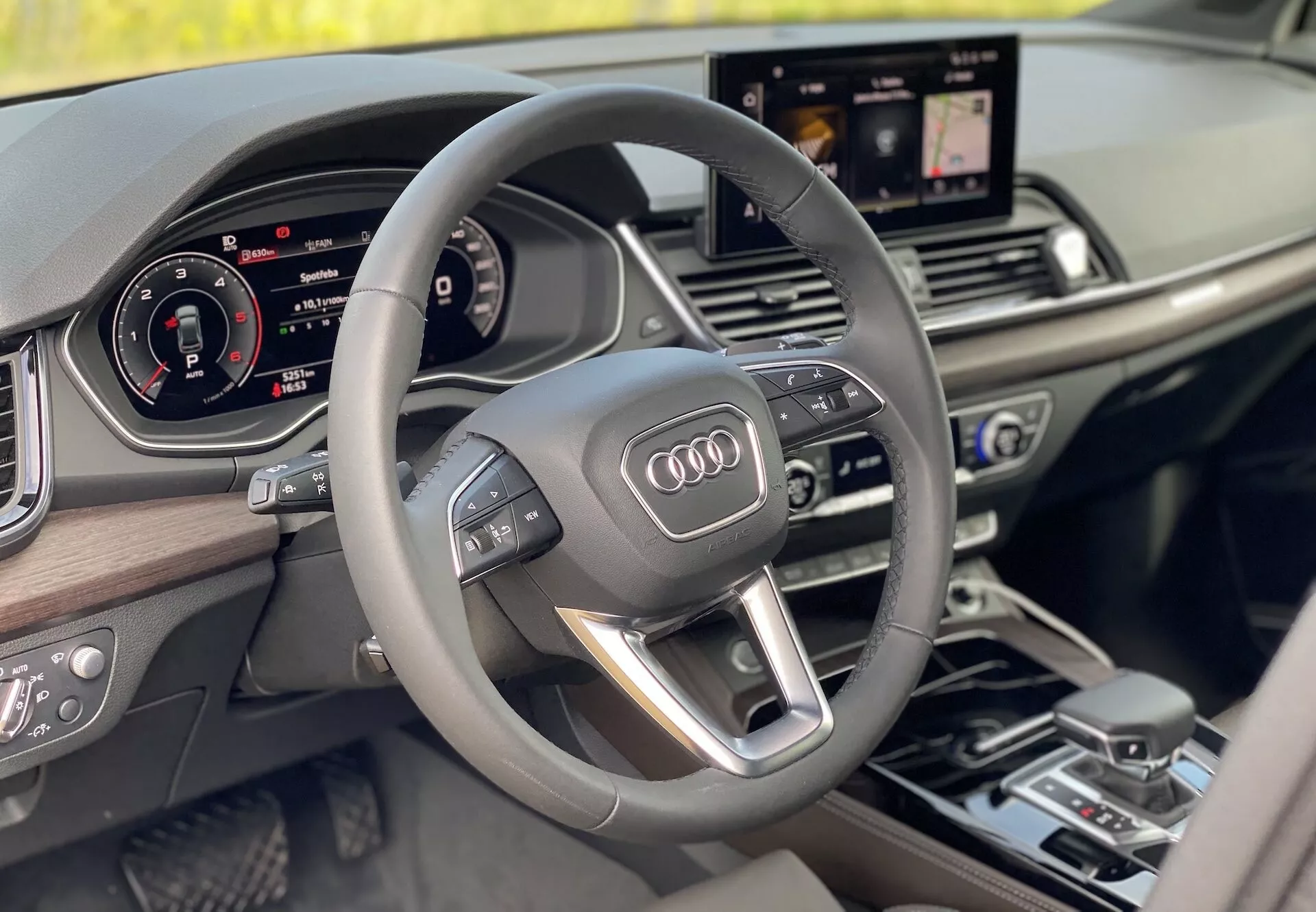 Lyxig interiör i Audi Q5 2020 Sportback ur förarens perspektiv, med en modern treekradig multifunktionsratt med integrerade kontroller, en helt digital instrumentpanel och en centralt placerad infotainmentskärm som visar navigering. Interiördesignen har trädetaljer som framhäver en känsla av lyx och modernitet.