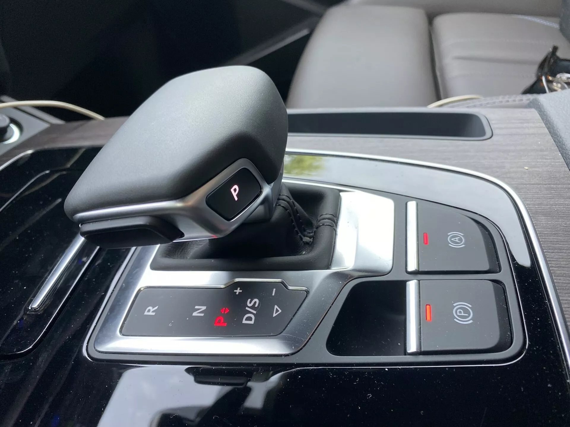 Närbild av växellägesväljaren för automatväxellådan i Audi Q5 2020 Sportback på mittkonsolen, med en shift-by-wire design i 'P' (Parkering) läget, alternativ för 'R' (Back), 'N' (Neutral) och 'D/S' (Körning/Sportläge), plus '+' och '-' symboler för manuell växling, en 'Auto Hold' knapp, och en elektronisk parkeringsbroms. Växellägesväljaren är omgiven av en blank svart panel och krom accenter, vilket indikerar ett lyxigt och tekniskt avancerat interiör.