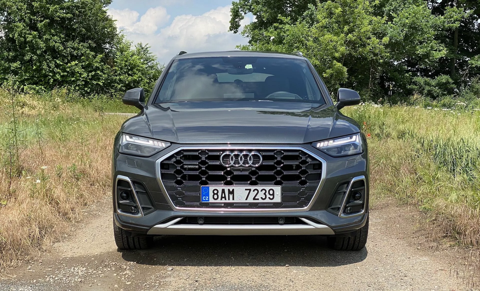 Frontal vy av en SUV Audi Q5 Sportback 2020 som visar en framträdande sexkantig Singleframe-grill med vertikala kromribbor, centralt placerad Audi-logotyp, skarpa Matrix LED-strålkastare med karakteristiska dagsljusljus, synliga luftintag med mönstrad design och främre stötfångare med integrerade dimljus och konturerade linjer. Bilen står på en grus- eller jordyta, i en naturlig, lantlig miljö med solljus som kastar skuggor, vilket framhäver den eleganta, sportiga looken och avancerade tekniska funktioner.