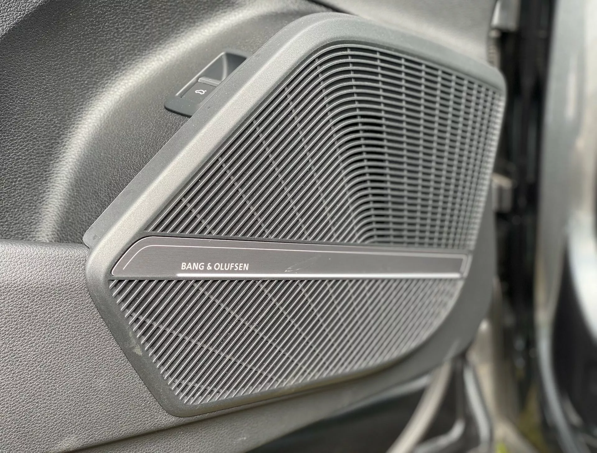 Närbild av ett premium BANG & OLUFSEN-högtalgaller på dörrpanelen i Audi Q5 Sportback 2020. Det fina nätmönstret på gallret är skarpt fokuserat, medan bakgrunden och förgrunden är suddiga. Ovanför gallret finns en liten triangulär sektion, som kan tänkas innehålla en diskanthögtalare, synlig från sidan och något framifrån. Bilden understryker fordonets ljudsystems lyxkvalitet.