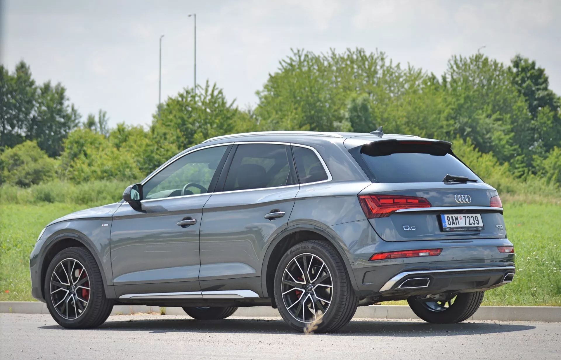 En grå Audi Q5 Sportback SUV från årsmodell 2020 visas i en tre-kvart bakifrån-vy som framhäver eleganta karosslinjer, flerspokedes aluminiumhjul, LED-bakljus, dubbla avgasrör och en sluttande bakre taklinje. Bilen står parkerad på en jämn yta med en naturlig bakgrund av gräs och träd, och det naturliga dagsljuset betonar dess sportiga designelement och kompakta utseende som en mellanstor SUV.