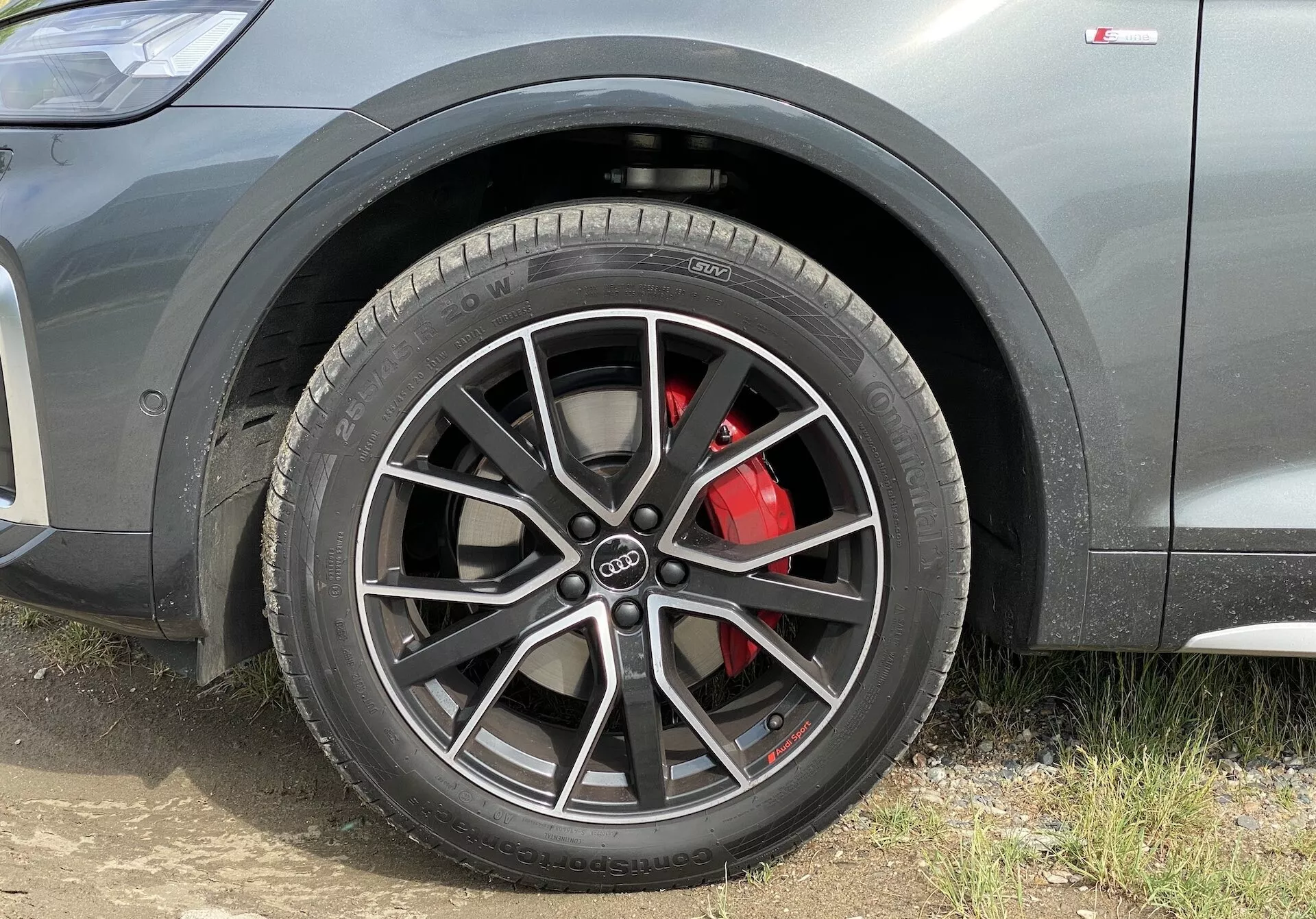 Närbild tagen från marknivå av det främre förarsidans hörnet på en Audi Q5 2020 Sportback. Fokuset ligger på ett flerspokat lättmetallhjul med centralt Audi-emblem, utrustat med en högpresterande lågprofildäck från Michelin Pilot. En röd bromsok syns bakom hjulet, vilket tyder på ett prestandaorienterat bromssystem. Fordonets karosseri är metallgrått med en synlig konturlinje och ett litet intag på sidokjolen, vilket betonar dess sportiga och moderna design.