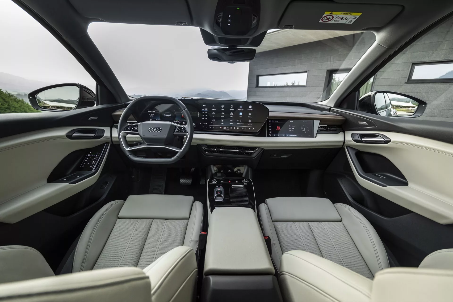Interiörvy av Audi Q6 e-tron 2024 från baksätets perspektiv med fokus på framsätena. De ljusa lädersätena fram har justerbara nackstöd och elektroniska justeringskontroller. Instrumentpanelen visar en bred digital mätarkluster och en stor central pekskärm för infotainmentsystemet, omgiven av kontrollknappar för klimat och körlägen. Mittkonsolen inkluderar mugghållare, förvaringsutrymme och en automatisk växelspak. Ratten är utrustad med flera kontroller och det panoramiska taket indikerar rikligt med naturligt ljus. Sammanfattningsvis framhäver bilden avancerad teknik och lyxiga fordonsfunktioner.