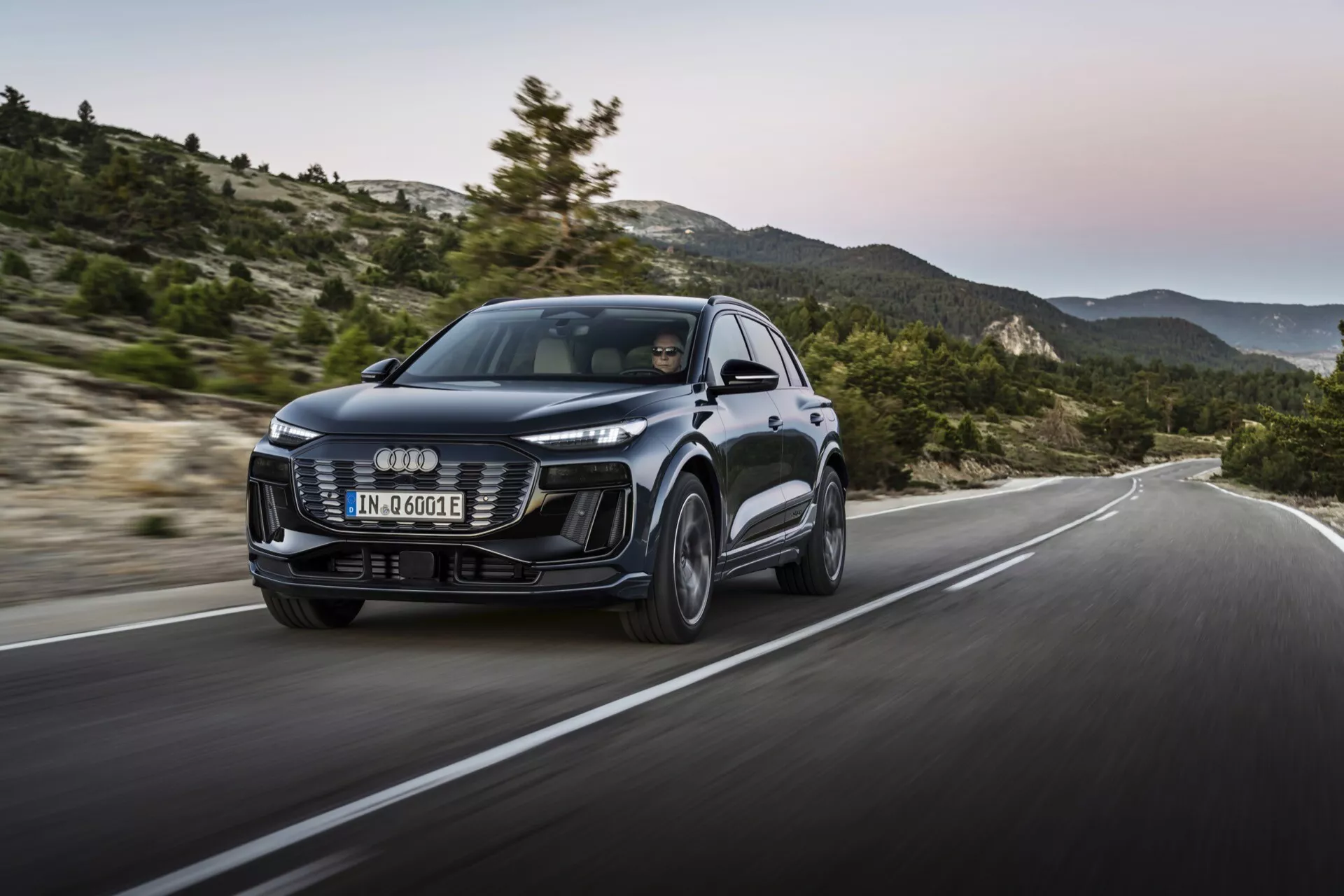 Audi Q6 e-tron 2024 med modern design, stor främre grill, eleganta LED-strålkastare och skulpturala linjer. Front-trekvartsvy med en dynamisk rörelsebild på en kuperad väg, som betonar prestanda och hantering.