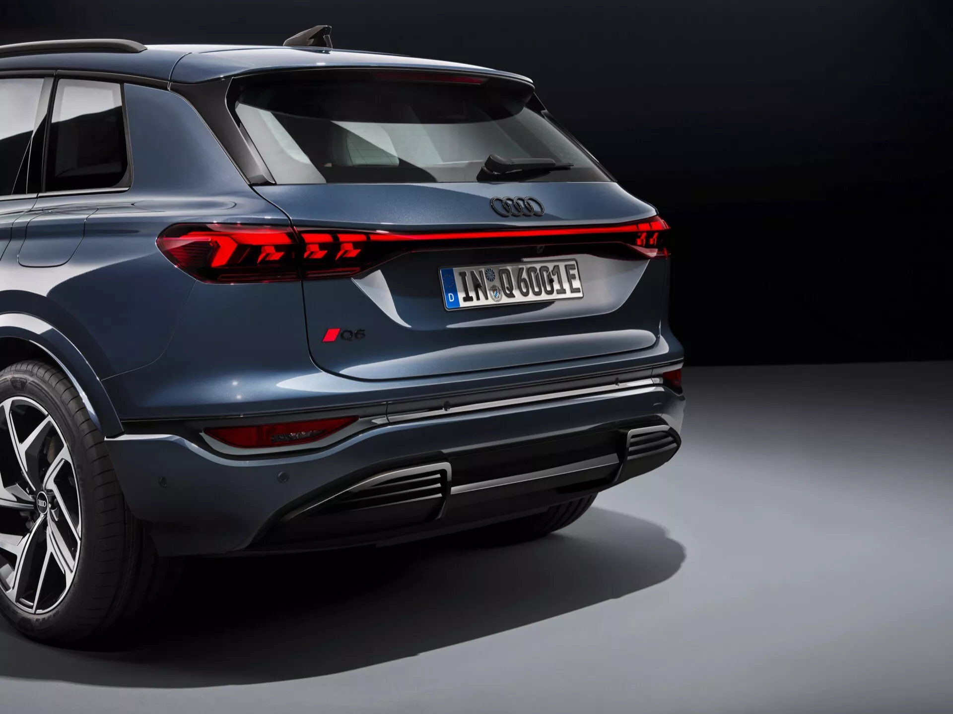 Bilden visar en modern Audi Q6 e-tron 2024 bil fångad från en bakre trekvartsvinkel som framhäver design- och teknikfunktioner. Den inkluderar en elegant, integrerad bakspoiler, en fullbredd bakre ljuslist med distinkta LED eller OLED signaturer, centralt emblem och kromlister som förbinder de bakre ljusen. Den bakre stötfångaren har parkeringssensorer, hjälpljus och en diffuser-stil design med framträdande utskärningar. Synliga sidoelement inkluderar ett stort fler-ekrade aluminiumhjul med högprestandadäck och en dynamisk fåra ovanför dörrhandtagen. 'e-tron' märket indikerar en elektrisk eller hybrid drivlina.