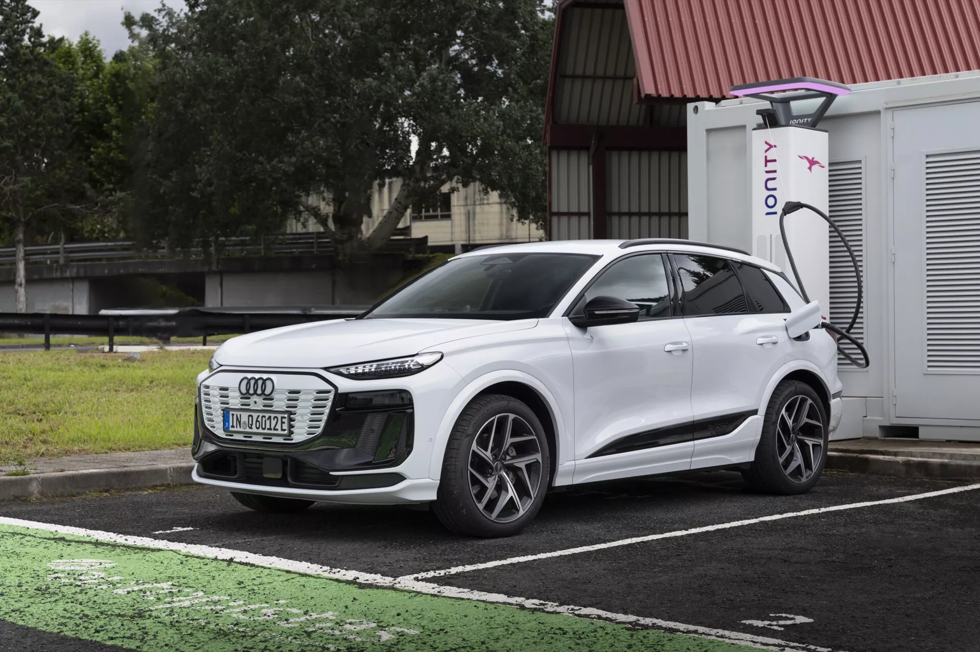 Trekvartsfrontvy av en vit 2024 Audi Q6 e-tron elektrisk SUV, som visar grillen fram, LED-strålkastarna, aerodynamiska linjer, multiekrade hjul och sidoprofilen. Fordonet är parkerat på en avsedd laddplats för elfordon med en synlig laddport och en IONITY-laddare i bakgrunden. Bilden kontrasterar bilens design mot en mörk, molnig himmel och våt trottoar, och illustrerar modern elfordonsteknik och avancerad belysning.