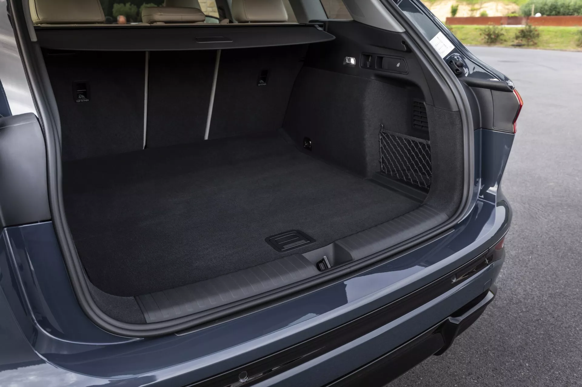 Bilden visar det öppna bagageutrymmet på en modern Audi Q6 e-tron 2024, fotograferad från en vinkel något ovanför och till höger om bilens bakre del. Föringsvinkeln erbjuder en klar syn på det rymliga, mattbeklädda lastutrymmet, som betonar dess djup och bredd. Noterbara funktioner inkluderar ett formgjutet fack till vänster för småsaker eller nödsituationer och en nätficka för förvaring till höger. En möjlig automatisk stängningsknapp är synlig på bagageluckan. Bakre stötfångaren och en delvis vy av högra bakljuset ger en vink om fordonets yttre design.