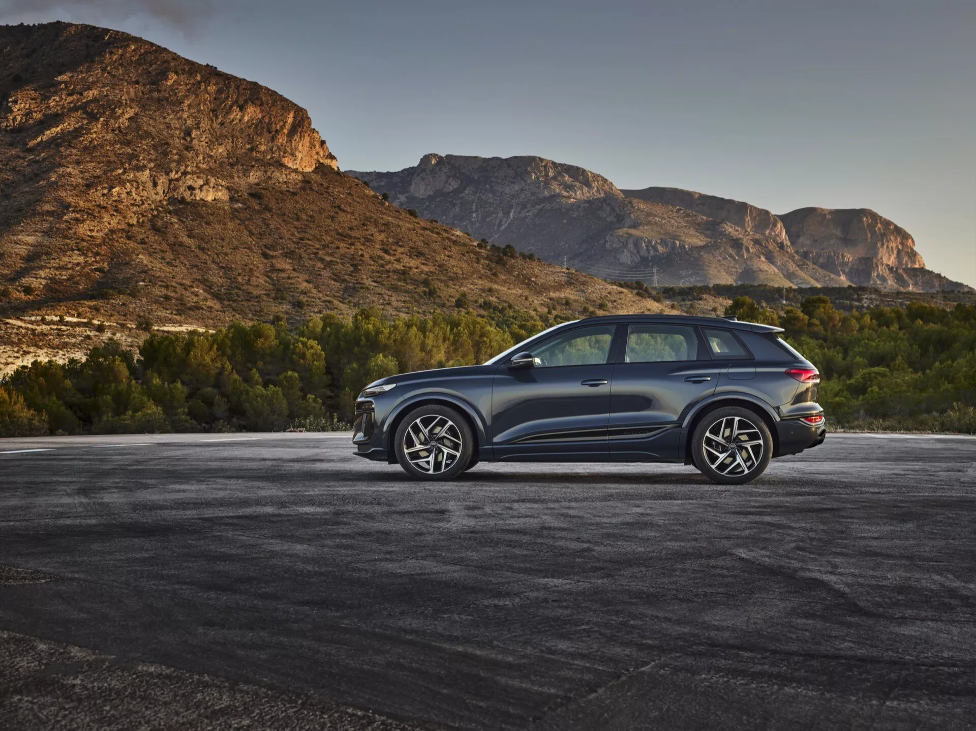 En modern SUV, trodd vara Audi Q6 e-tron 2024, parkerad utomhus med en bergig bakgrund, fångad i trekvarts bakifrån från en upphöjd vinkel. Karossens mörka metalliska färg kontrasterar med den naturliga miljön, vilket framhäver eleganta linjer och aerodynamiska konturer. Synliga detaljer inkluderar LED-bakljus, stora lättmetallfälgar med flerspokedesign, dubbelt avgasrörssystem, tonade rutor, panoramasoltak och skyddande lister längs de nedre kanterna. Bakrutan är lite lutad med en bakspoiler och stora sidospeglar som troligen har integrerade blinkers, vilket ökar fordonets sportiga och lyxiga utseende.