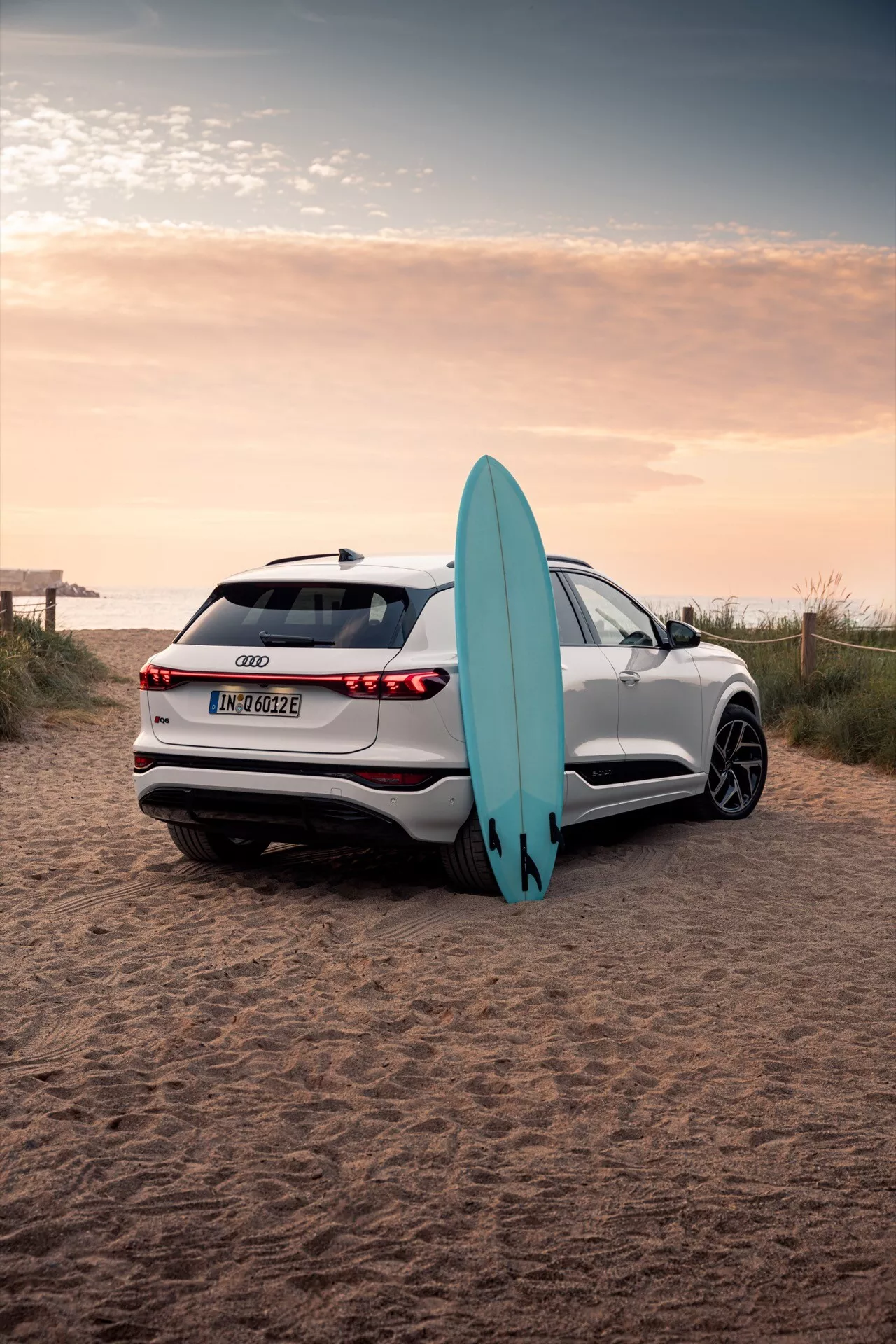 Bilden visar en vit Audi Q6 e-tron 2024 SUV parkerad på en sandig strandmiljö med en surfbräda. Fotograferad från en bakre trekvartsvinkel fångas bilens eleganta linjer, LED-bakljus, parkeringssensorer, backkamera och flerspetsiga lättmetallfälgar. Solnedgångens reflektion förstärker den avancerade lackkvaliteten, medan surfbrädan lutande mot bilen betonar ett äventyrligt livsstilselement.