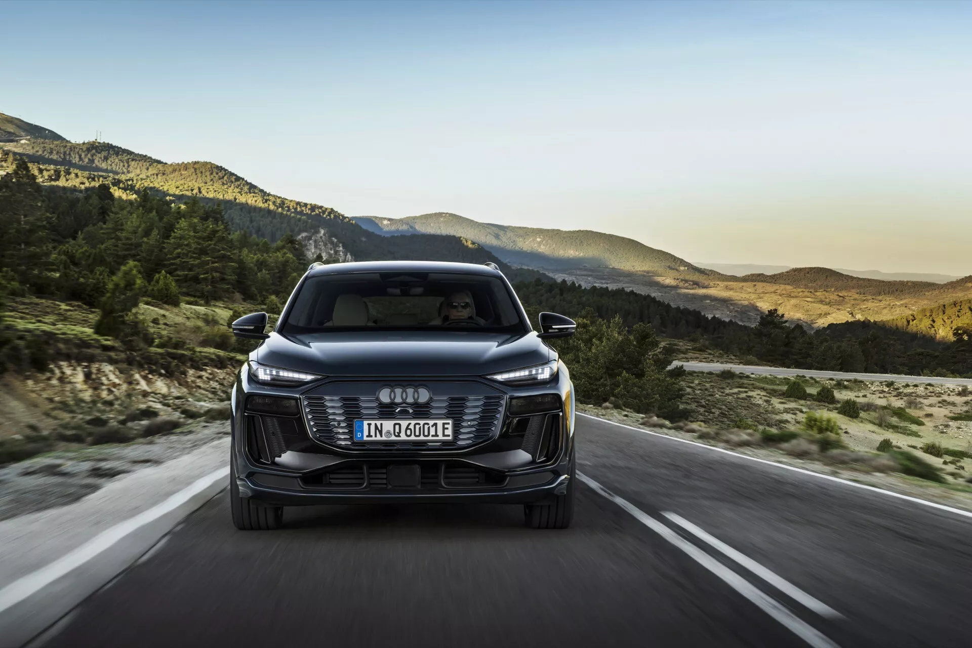 Frontal vy av 2024 Audi Q6 e-tron, som visar dess frontgrill, strålkastare och stötfångare med de fyra sammankopplade ringarna som logo. Den europeiska registreringsskylten är monterad på grillen, och beteckningen 'e-tron' indikerar dess elektriska/hybrida natur. Bilen kör på en öppen väg med ett bergigt landskap i bakgrunden, vilket antyder en sen eftermiddagmiljö. Funktioner inkluderar eleganta LED-strålkastare, estetisk grill, förarassisterande sensorer/kameror, aerodynamiska speglar och en effektiv design.