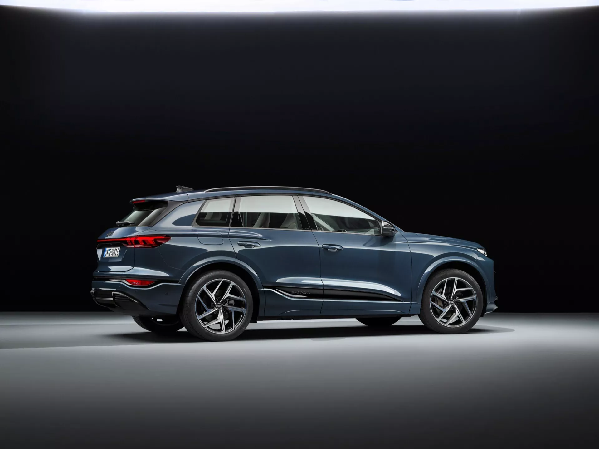 Lågprofilvinkelbild av 2024 Audi Q6 e-tron i en studiouppsättning med mörk bakgrund, med sportiga mångekrade lättmetallfälgar, flytande karossdesign med karaktärslinje, aerodynamiska sidospeglar med integrerade blinkers, kromade fönsterramar, panoramasoltak, takräcken, måttlig markfrigång och infälld dubbla avgasrörssystem.