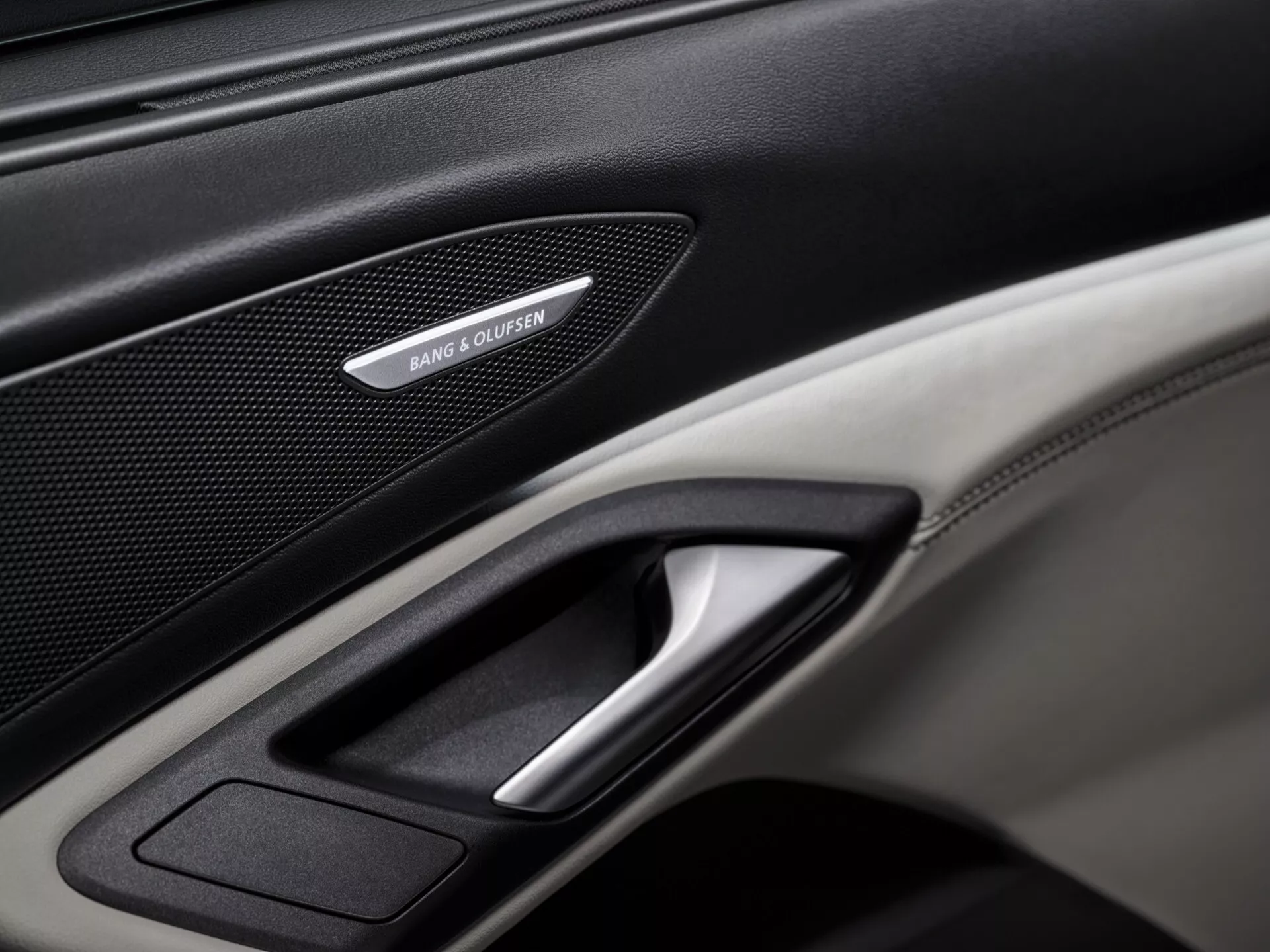 Närbild av Audi Q6 e-tron 2024 GF:s inre dörrpanel, som framhäver premiumfunktioner såsom ett Bang & Olufsen-högtalargaller, en kromad dörrhandtag och en kombination av läder och kolfibertexturer. Den väl upplysta bilden fokuserar på de intrikata detaljerna i panelen och visar fordonets lyx- och teknologielement.