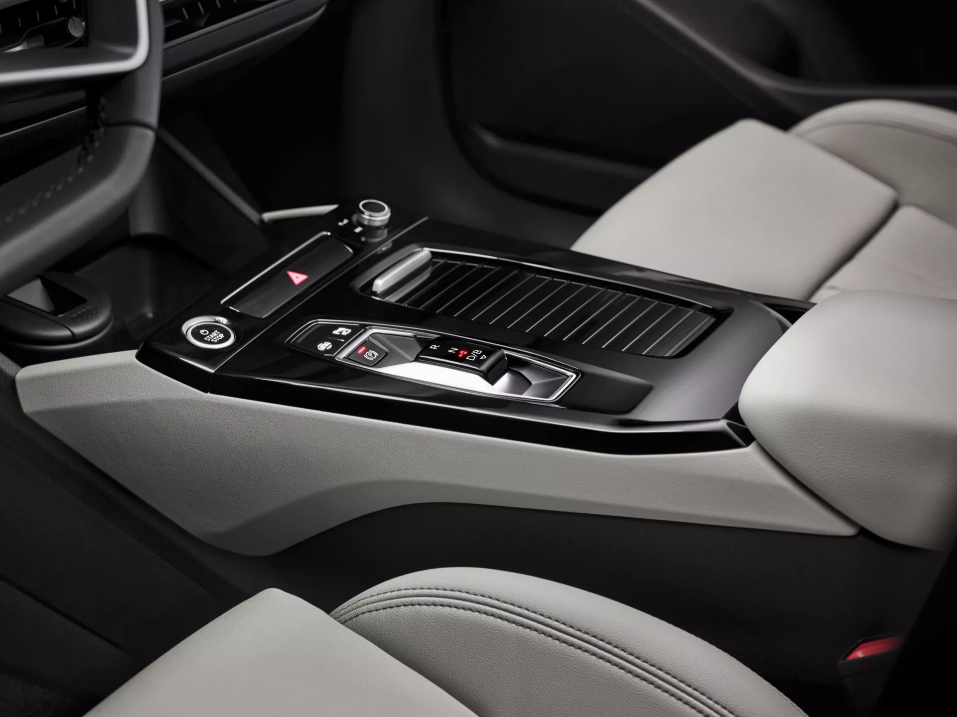 Interiörbild av Audi Q6 e-Tron 2024 med fokus på mittkonsolområdet. Bilden, tagen ur passagerarperspektiv, framhäver en blank svart växelspak med metalliska detaljer, flera kontrollknappar, ett litet förvaringsutrymme med skjutlock, två mugghållare, en läderklädd armstöd, klimatkontrollventiler och delar av ljusa lädersäten med mörkare kontrasterande sektioner. Mittkonsolens funktioner visar en modern design och lyxiga element.