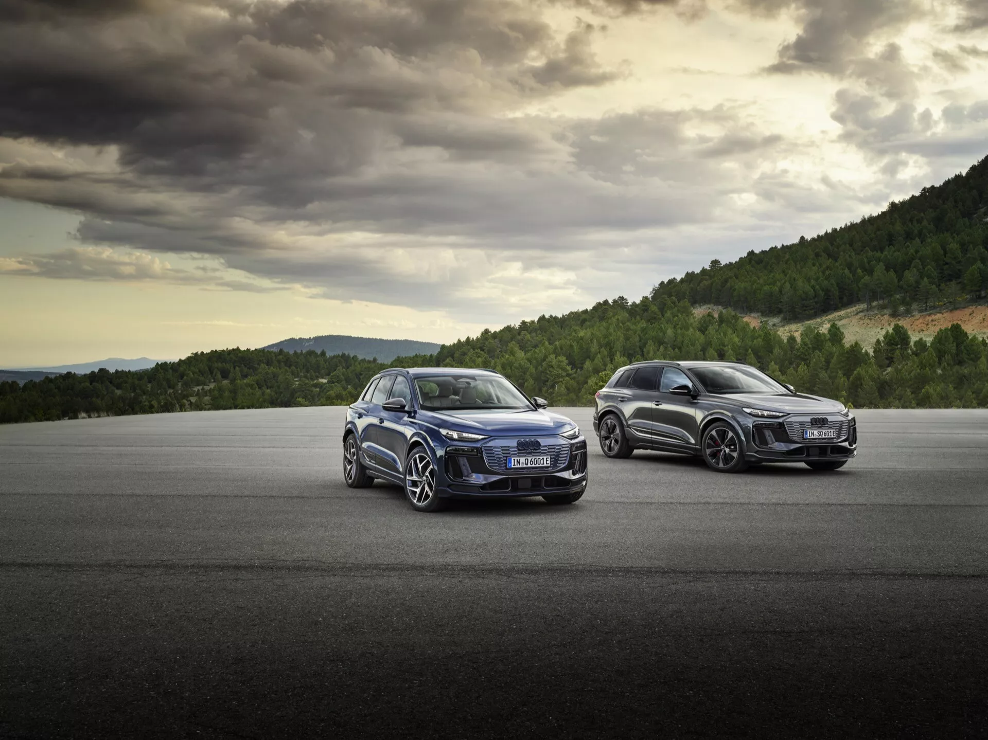 Bilden visar två moderna SUV:ar, sannolikt från samma tillverkare, placerade på en asfaltyta med ett naturlandskap i bakgrunden under en molnig himmel. I förgrunden finns en sportig bil som visar sin vänstra främre kvartal, inklusive grillen, strålkastarna, det vänstra framhjulet och dörren, en del av vindrutan och huven, vilket framhäver framträdande veck på huven och sofistikerade LED-strålkastare. Den andra bilen, något bakom, är sedd från sin högra bakre kvartal och visar passagerarsidans bakljus, en del av bakre stötfångaren och bakhjulen. Den låga vinkeln betonar fordonens hållning och designdetaljer och antyder lyx och prestanda med stora grillar och högkvalitativa fälgar, med fokus på exteriör estetik och moderna bilteknologier.