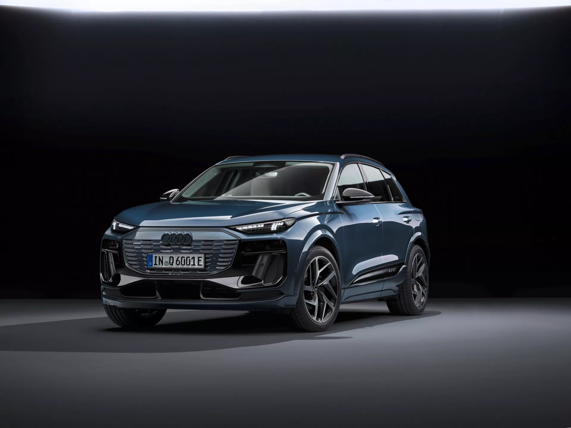 Bilden visar en modern Audi Q6-e-tron 2024 SUV från en trekvarts frontvy som framhäver både dess front- och sidoprofil. Viktiga identifierare inkluderar Audi-badgen med fyra ringar på grillen och e-tron-badgen som indikerar dess elektriska natur. Bilden är tagen från ett lågt perspektiv, vilket förstärker fordonets resning och visar dess designlinjer, muskulösa hjulhus och aerodynamiska funktioner. Bilen är centrerad i ramen, placerad på en slät reflekterande yta med mjuk, diffus belysning och en mörk gradering i bakgrunden som betonar fordonets design. Synliga tekniska funktioner inkluderar LED-strålkastare, dagsljuslampor (DRL), en digital display och legeringshjul med en komplex design. Den övergripande polerade presentationen antyder en reklamfotografi som framhäver fordonets teknik, design och lyxaspekter.