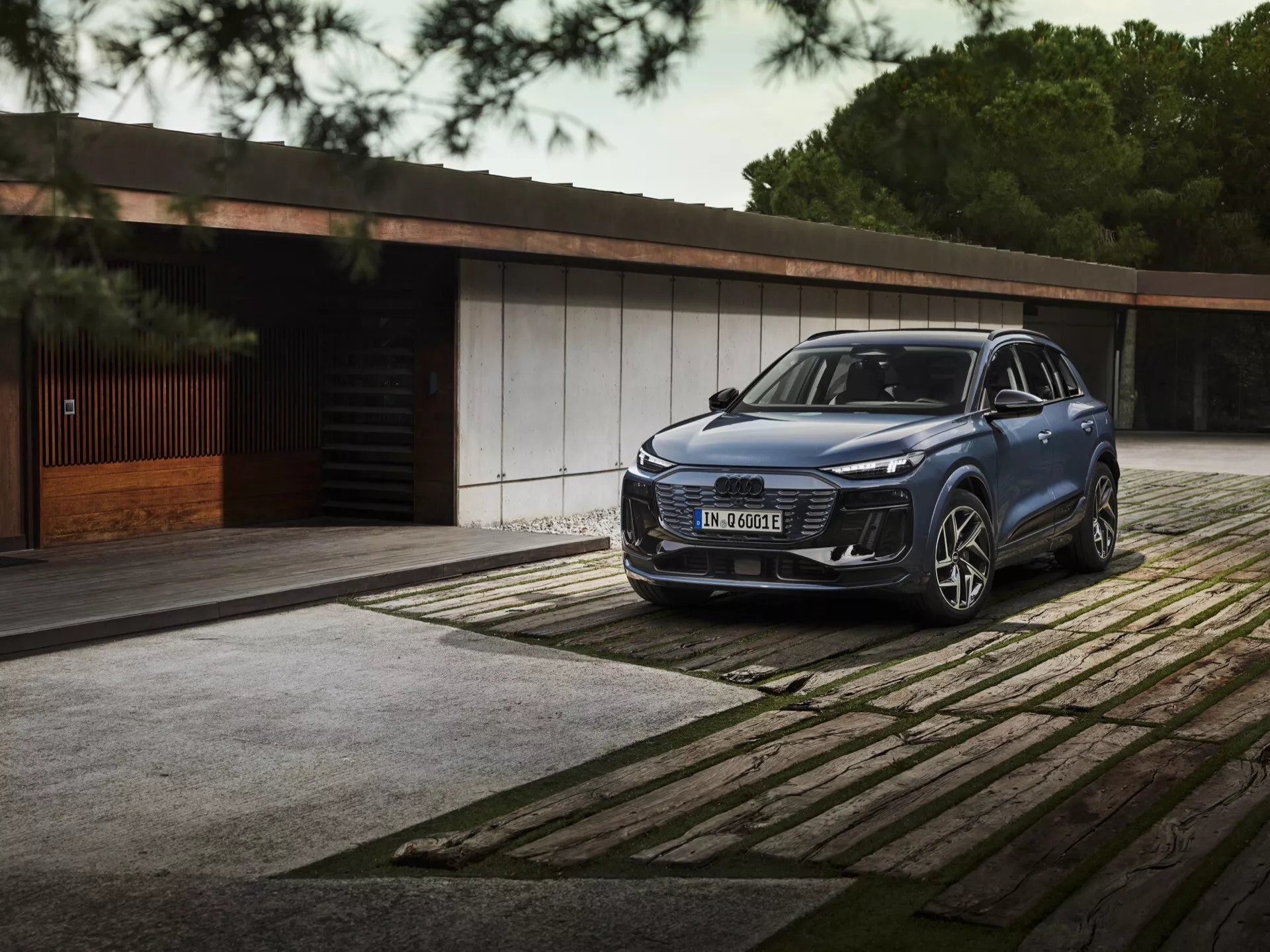 2024 Audi Q6 E-Tron tre kvarts främre vy SUV med eleganta LED-strålkastare, framträdande grill och stort emblem parkerad på mönstrad trottoar. Lyxig byggnad och träd i bakgrunden betonar lyx och exklusivitet.